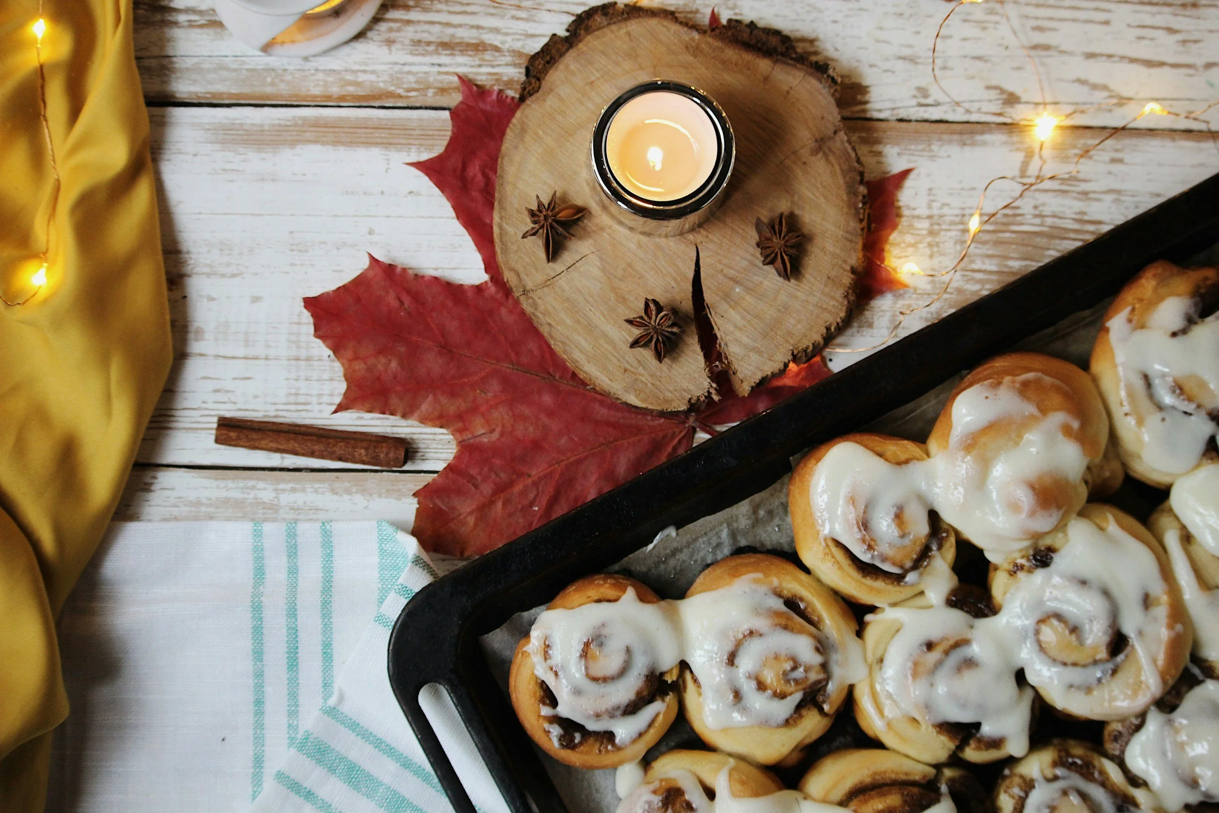 A tray of cinnamon rolls with icing on a baking sheet on a white wooden table, with autumn leaves, a candle, and star anise around it.