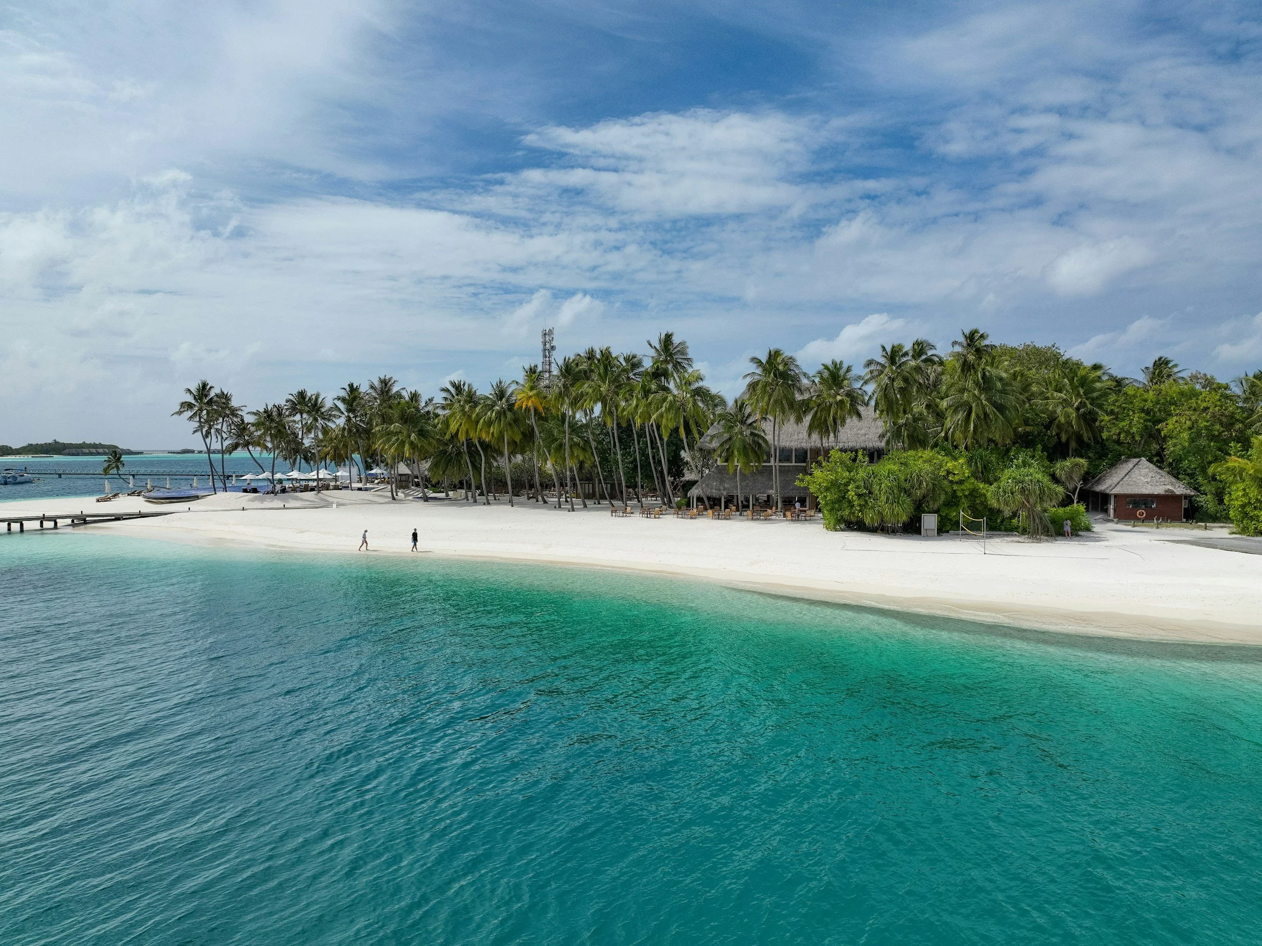 Tropical beach with white sand, emerald water, and numerous palm trees behind beachside huts under a partly cloudy sky.