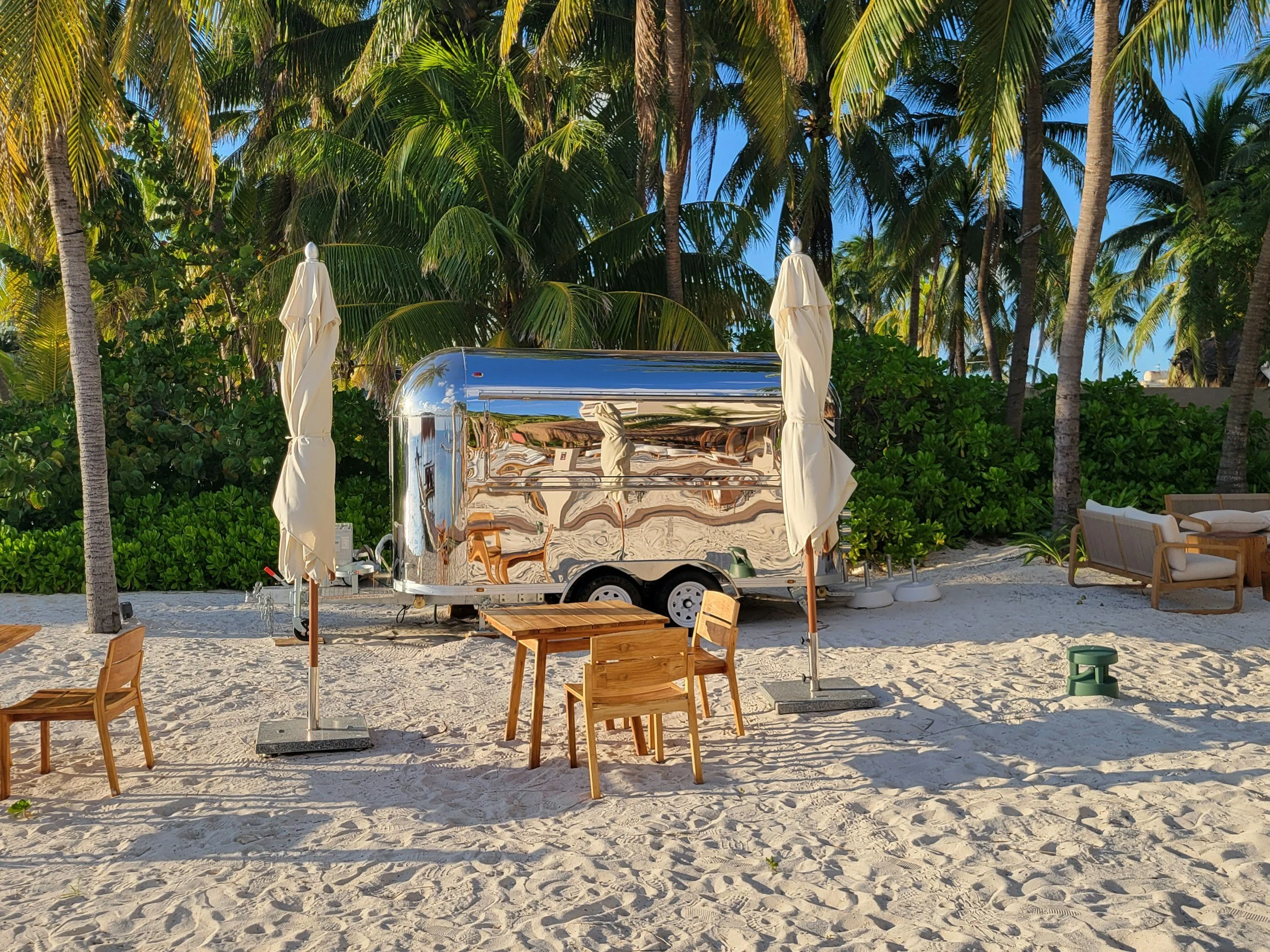 A shiny silver food truck or trailer on a sandy beach, surrounded by tropical trees and outdoor furniture such as chairs, tables, and umbrellas.