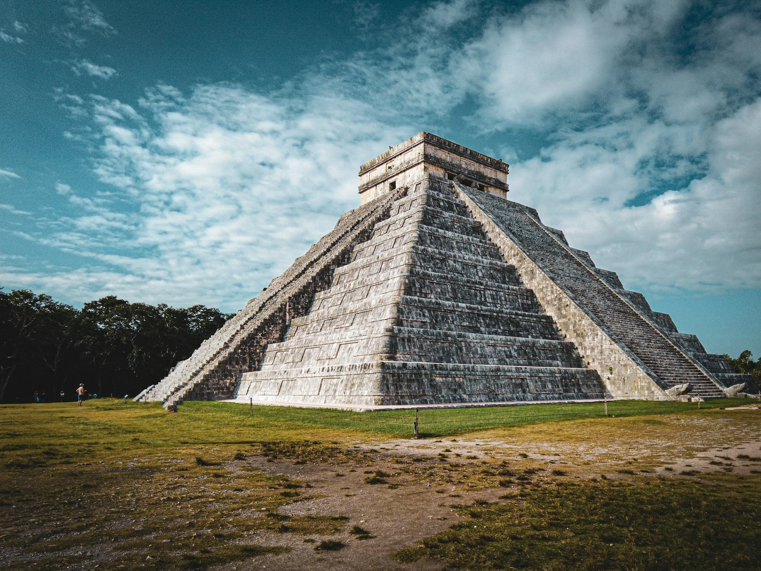 The ancient Mayan pyramid of El Castillo at Chichen Itza under a partly cloudy sky, surrounded by grassy grounds and trees in the distance.