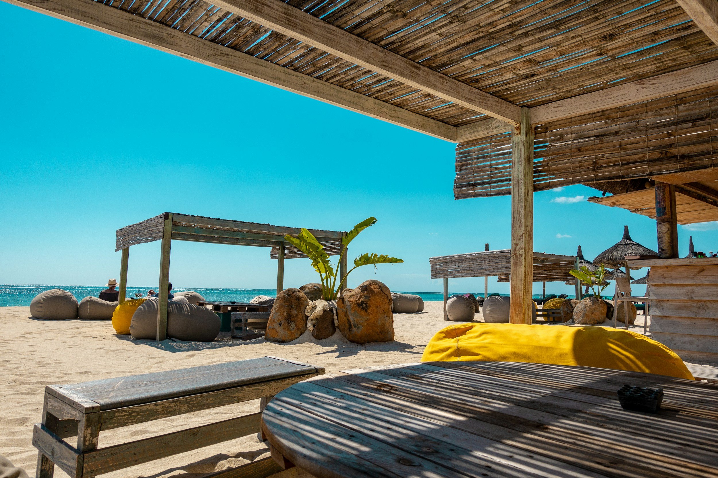 Beachside hut with wooden furniture, bean bag chairs, sun umbrellas, and a view of the ocean under a clear blue sky.