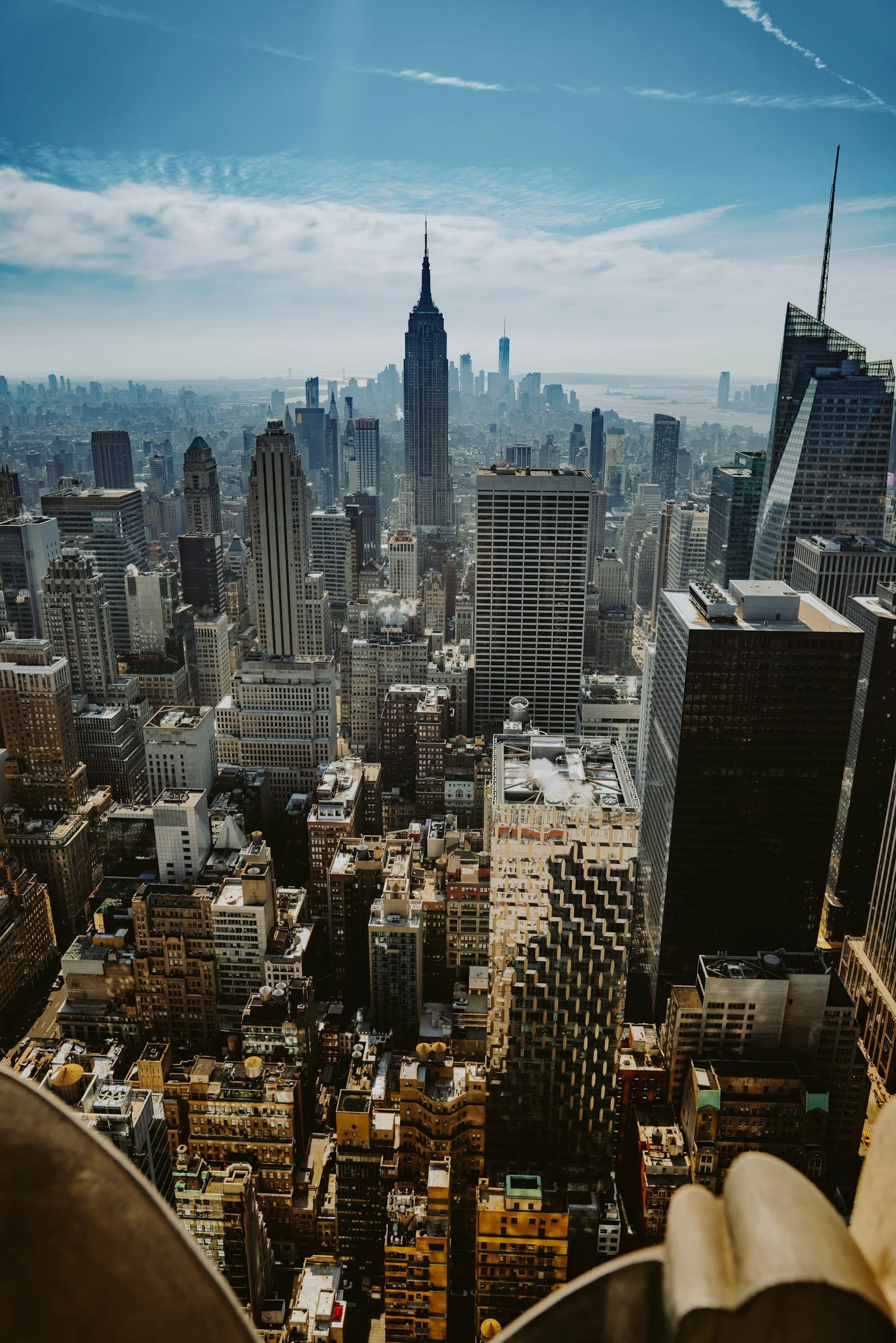 Aerial view of New York City skyline with the Empire State Building in the center and One World Trade Center in the distance under partly cloudy blue sky.