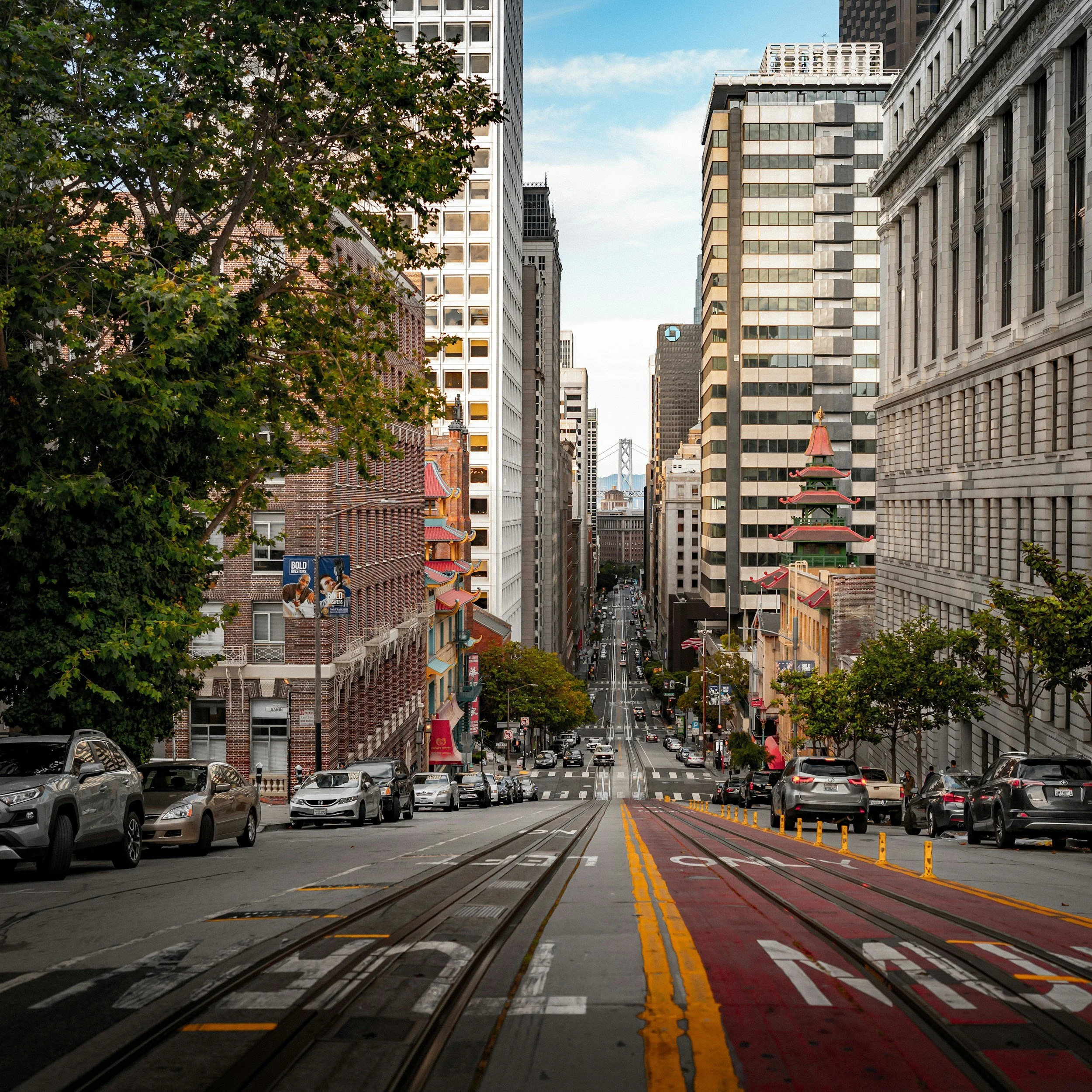 City street with cars parked along the curb, subway tracks running down the center, and tall buildings on either side under a partly cloudy sky.