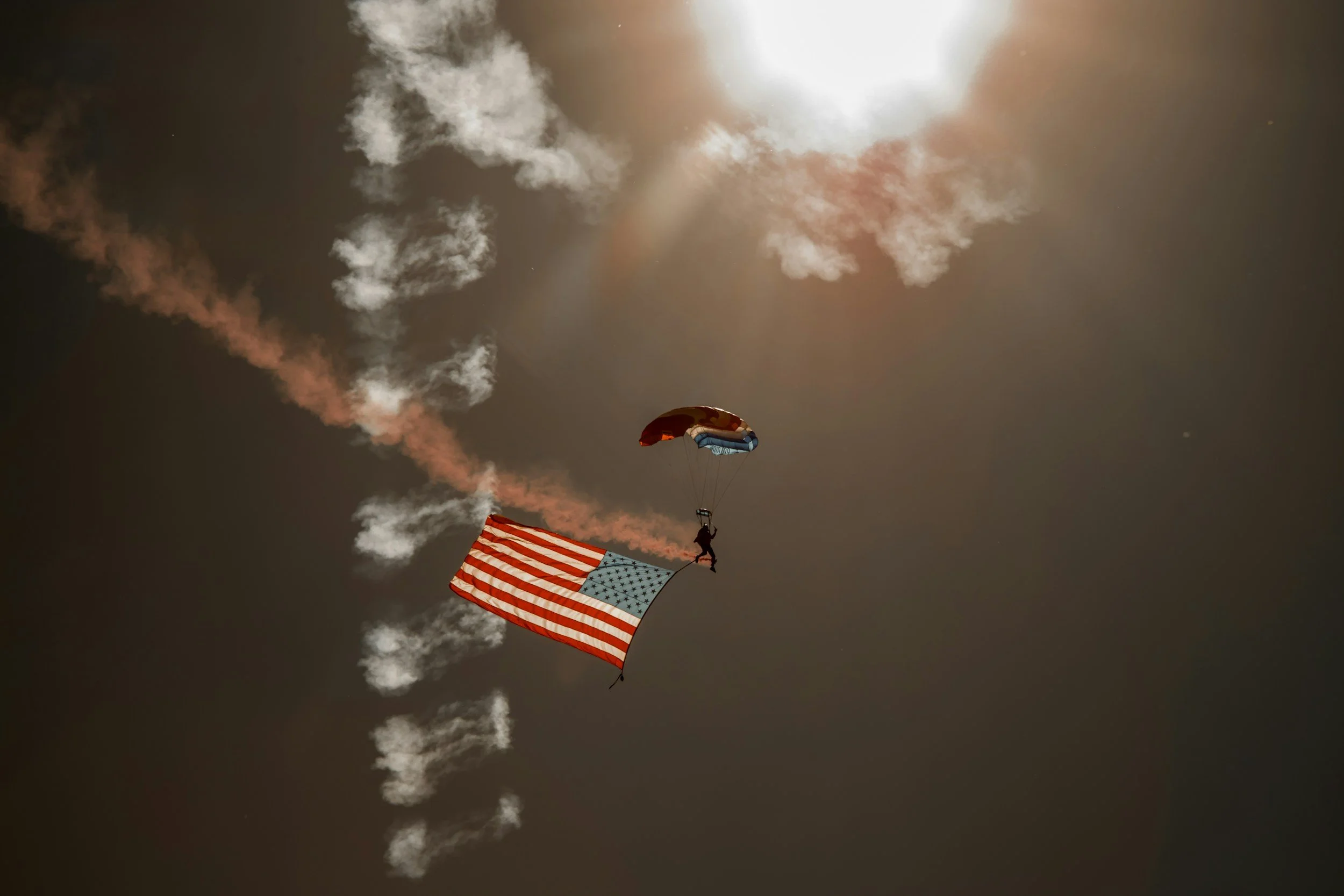 Skydiver descending with an American flag, paratrooper with a red, white, and blue parachute, against a cloudy sky with the sun shining.