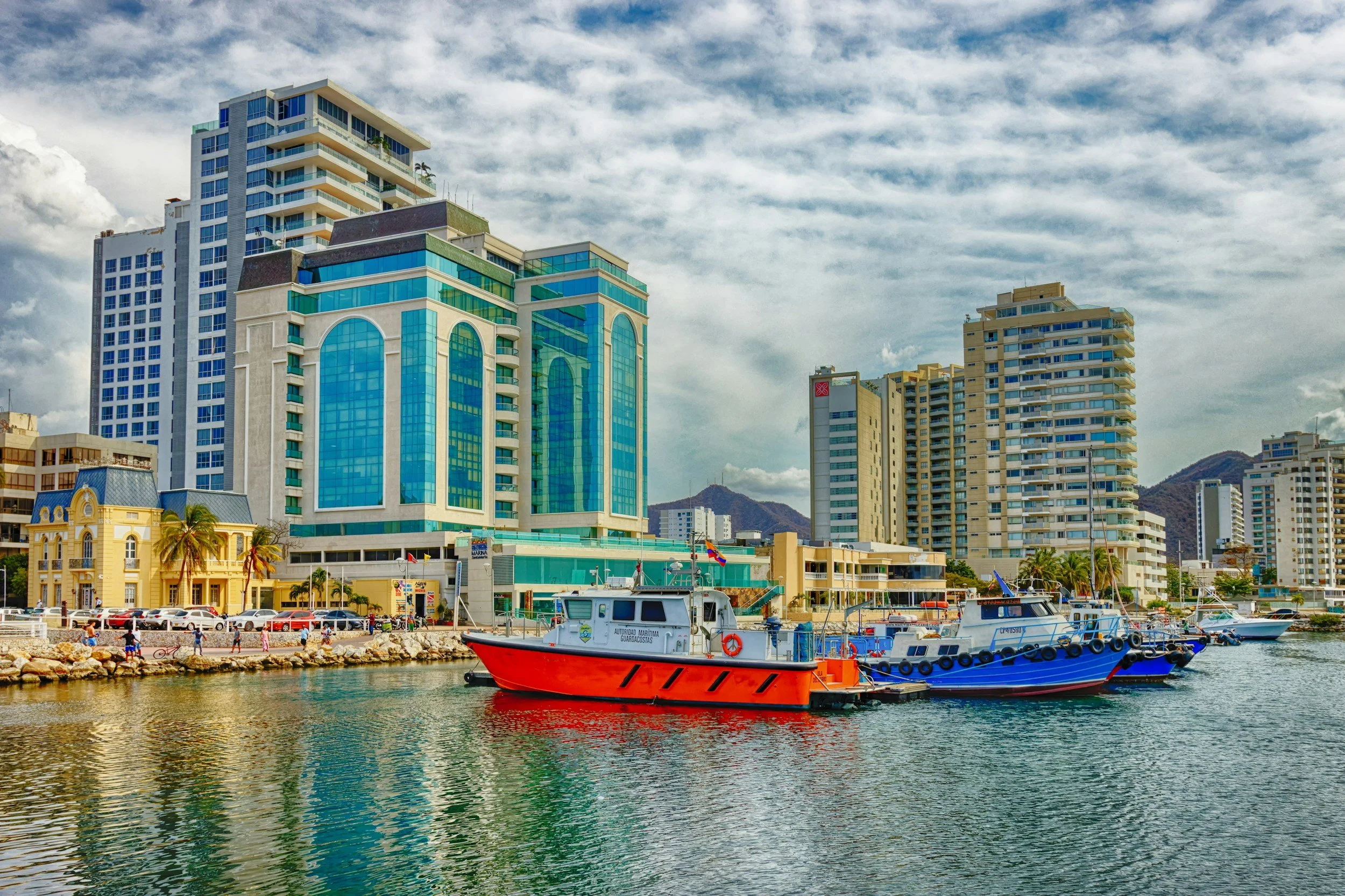 Cityscape with modern high-rise buildings and boats docked along the harbor, under a cloudy sky.
