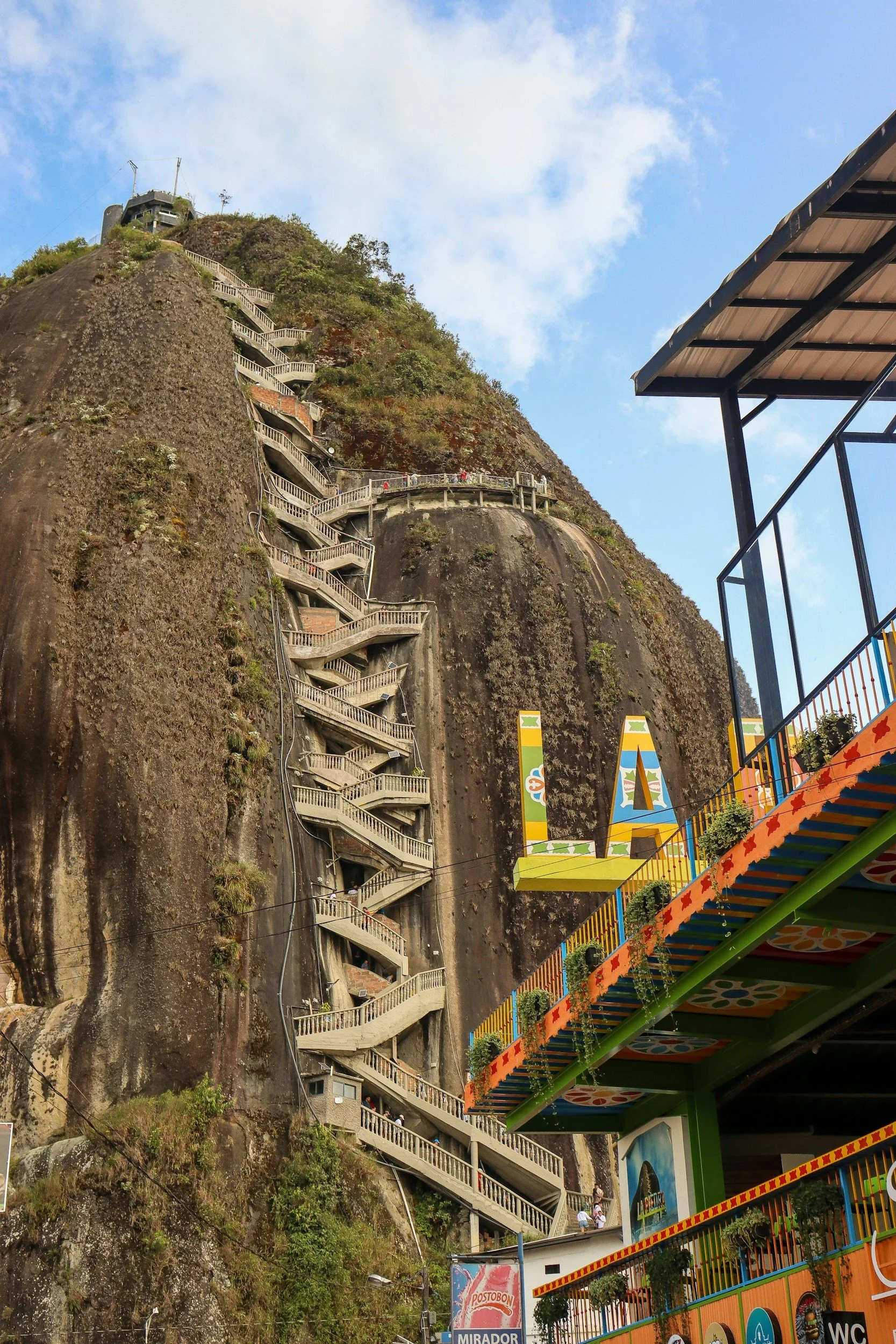 A rocky hillside with a zigzagging staircase built into it, leading to a structure at the top. In the foreground, there's a colorful building with signage for 'L.A.' and various decorations.