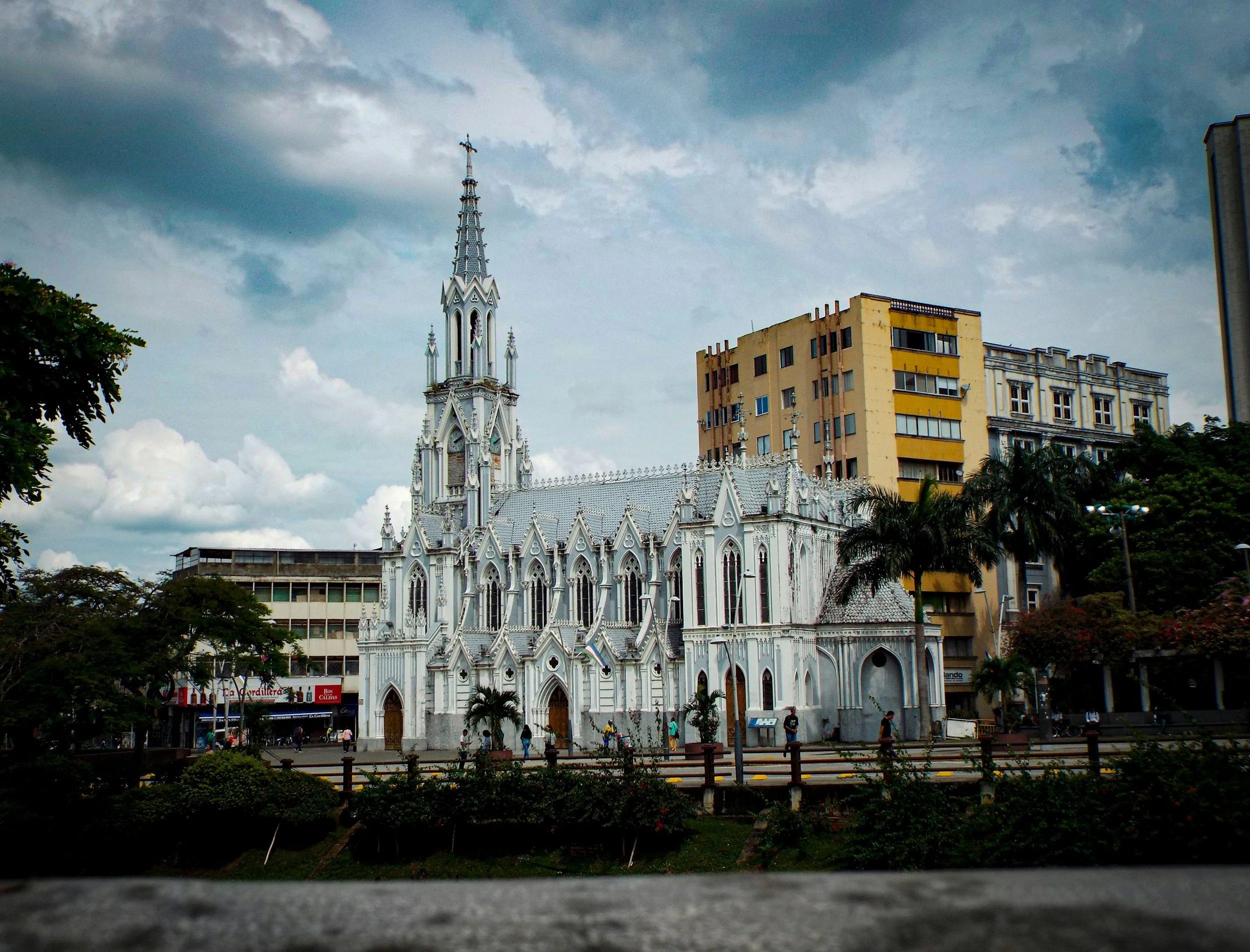A white Gothic-style church with a tall spire, surrounded by modern and historic buildings, with trees and a cloudy sky in the background.