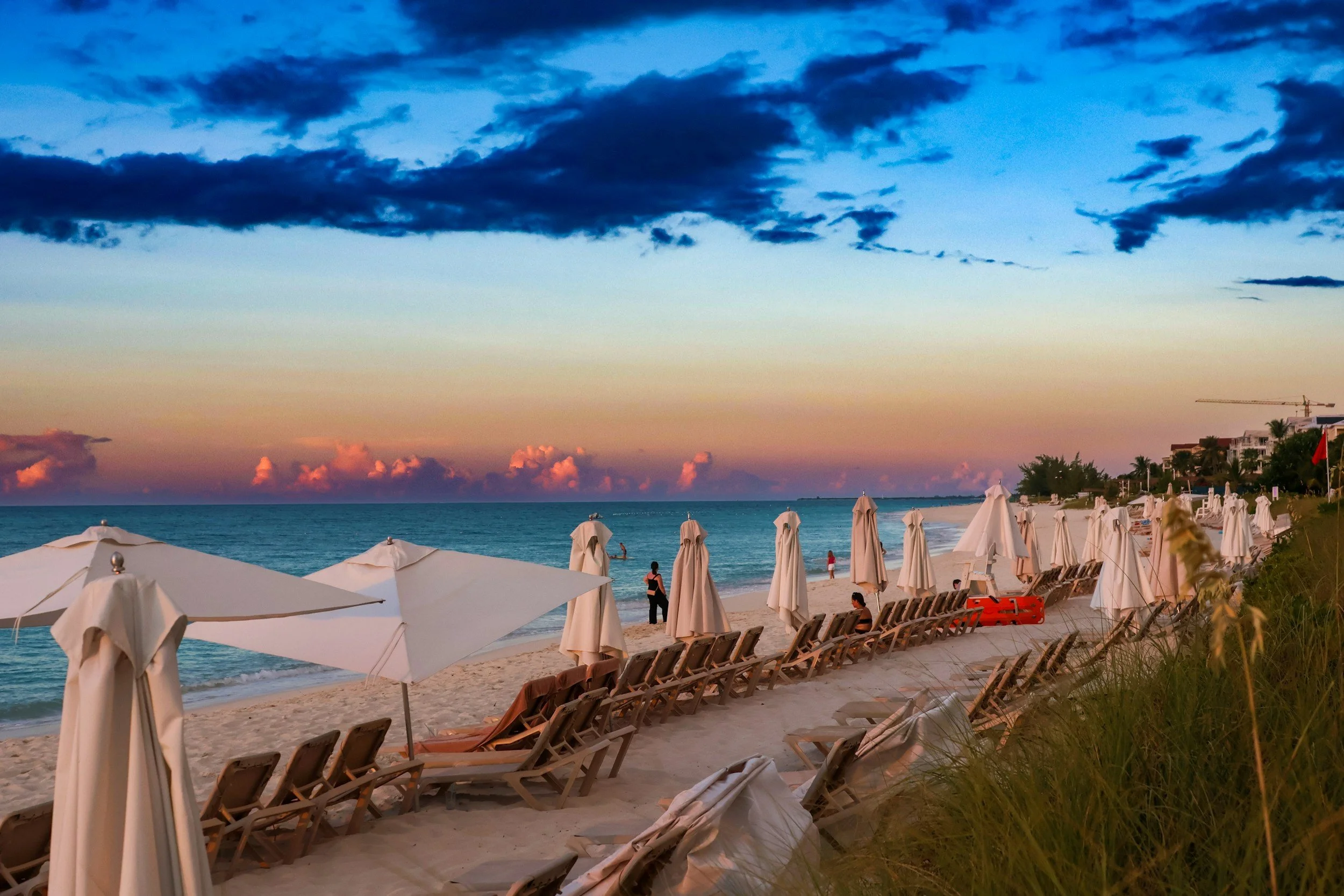 Empty beach with rows of white umbrellas and lounge chairs facing the ocean during sunset, with a cloudy sky and some people in the distance.