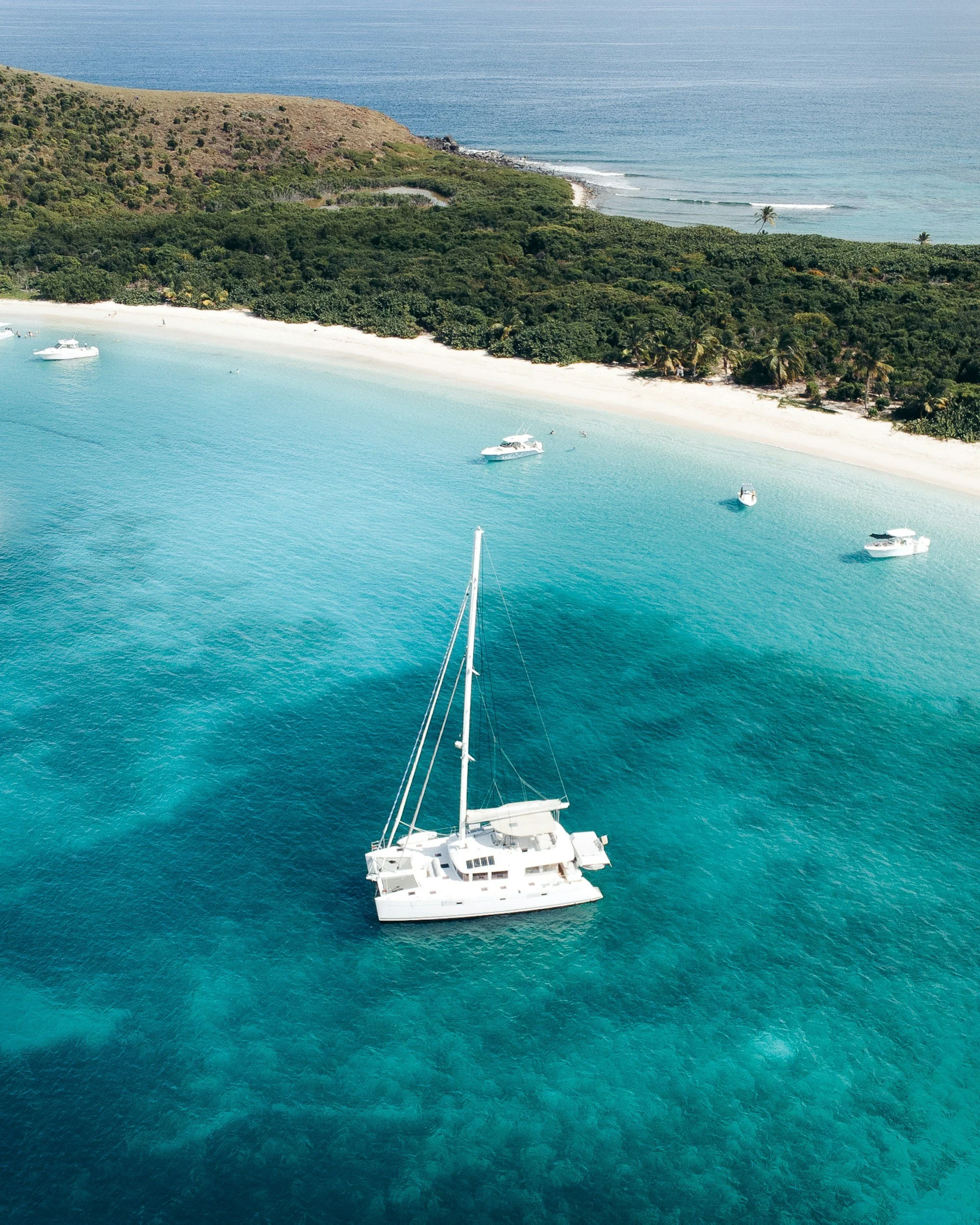 Aerial view of a white yacht floating in clear turquoise ocean near a tropical island with lush green foliage and a sandy beach.