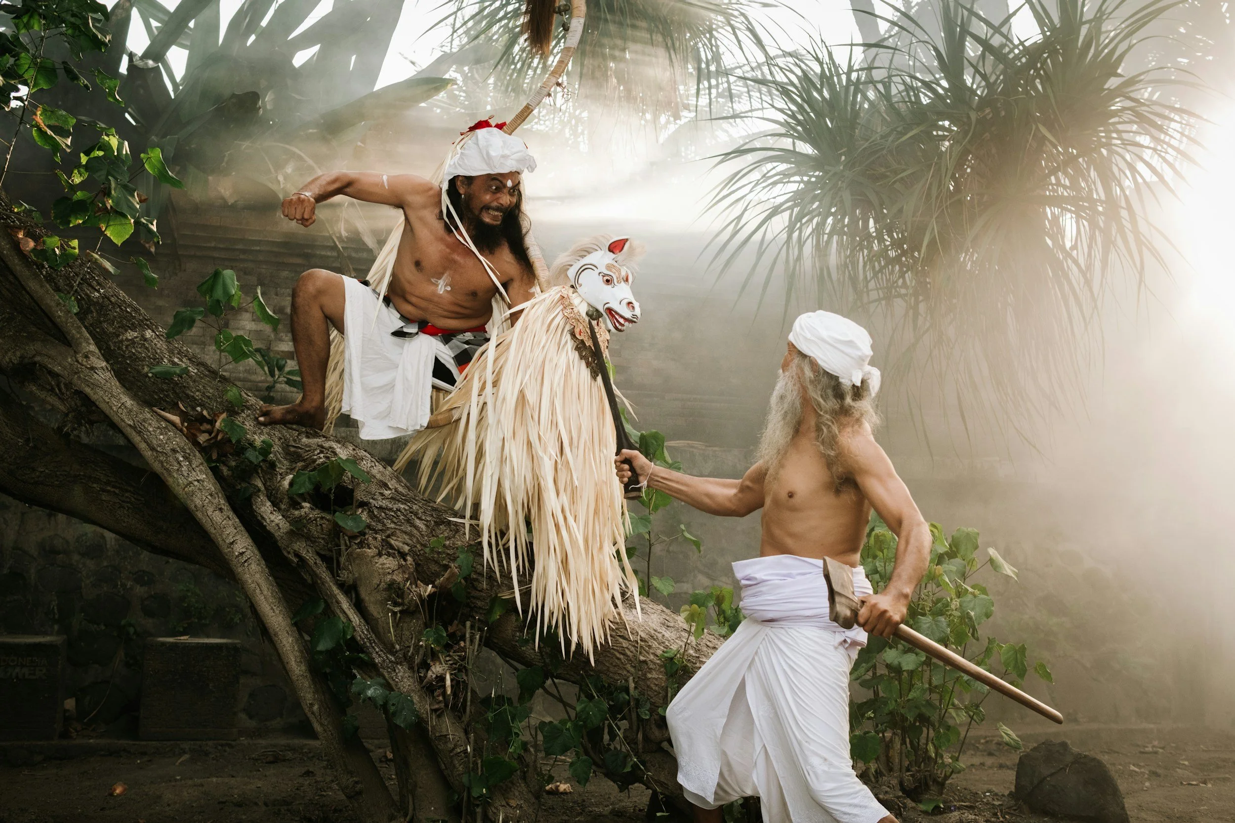 Two men with long white hair and beards, dressed in traditional white garments, engage in a puppet show with a large, elaborate puppet and a man in tribal attire on a tree branch in a jungle setting.