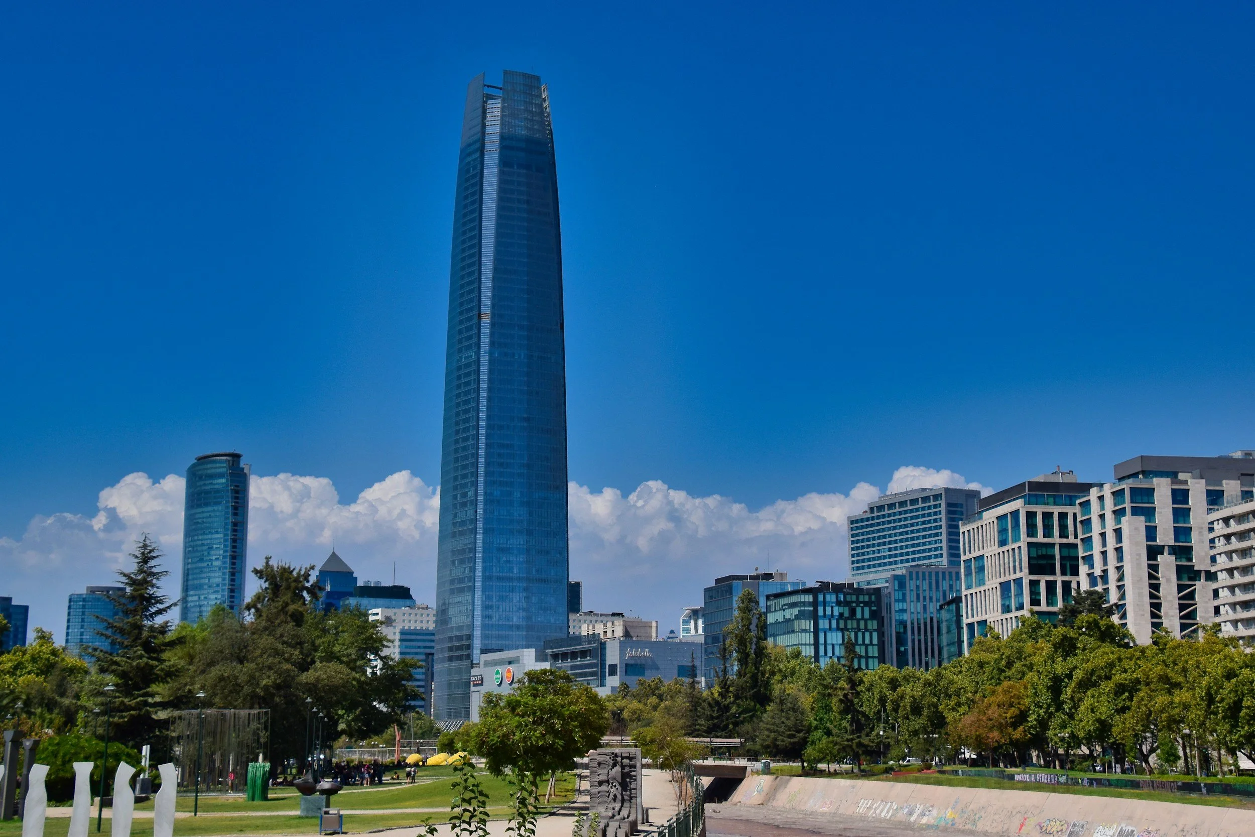 Skyscrapers in downtown San Francisco, including the Salesforce Tower, with green park trees in the foreground and a blue sky with clouds.