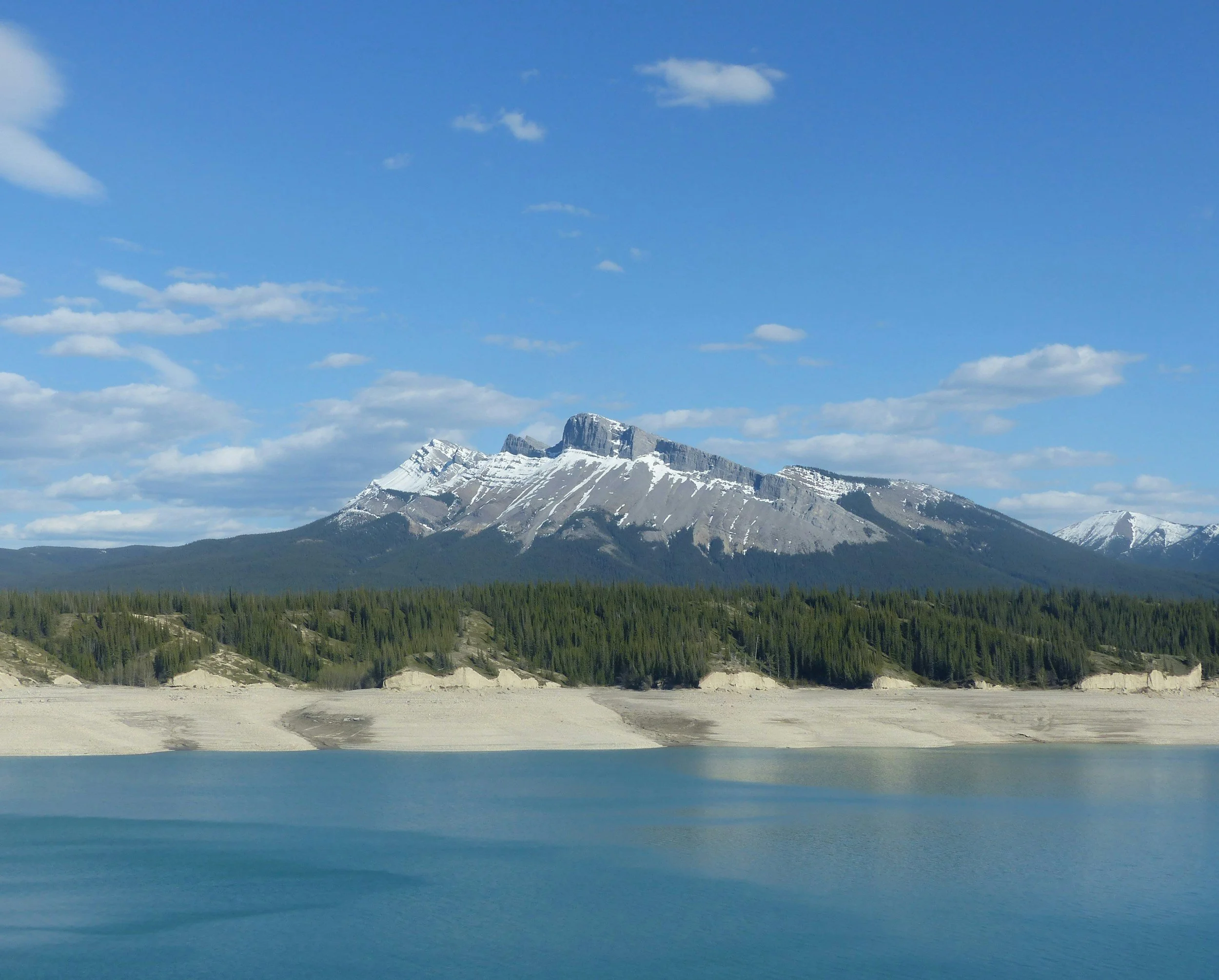 A serene mountain landscape with snow-capped peaks, a dense forest of pine trees, and a calm lake reflecting the scenery under a partly cloudy blue sky.