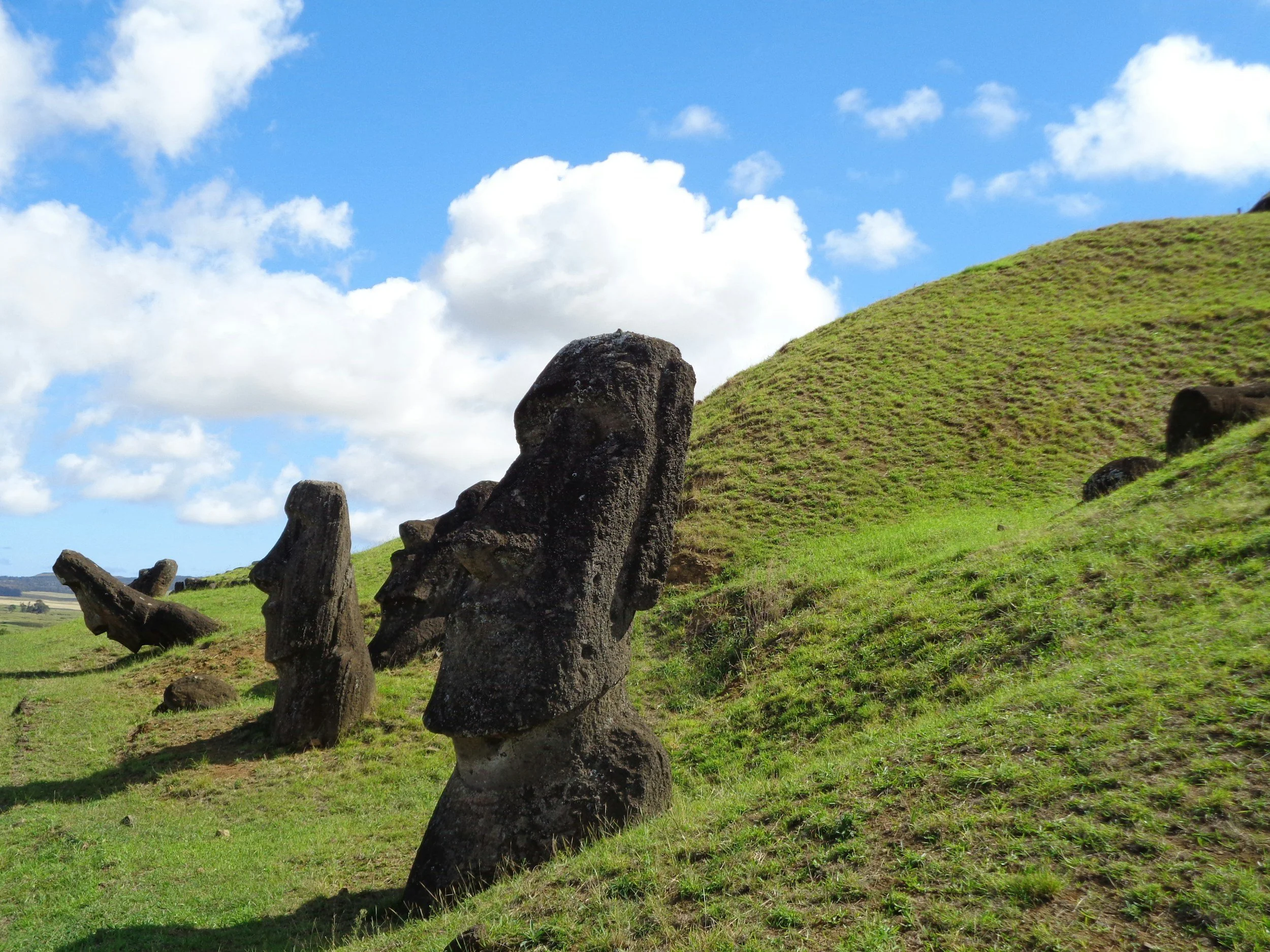Several moai statues on a grassy hillside under a partly cloudy sky, with a green hill in the background.