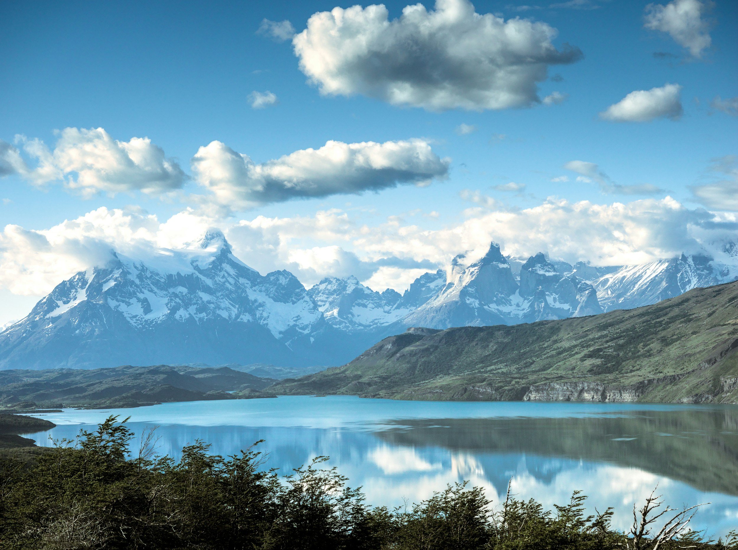 Snow-capped mountains in the background with a large lake reflecting the mountains and sky, and green vegetation in the foreground.