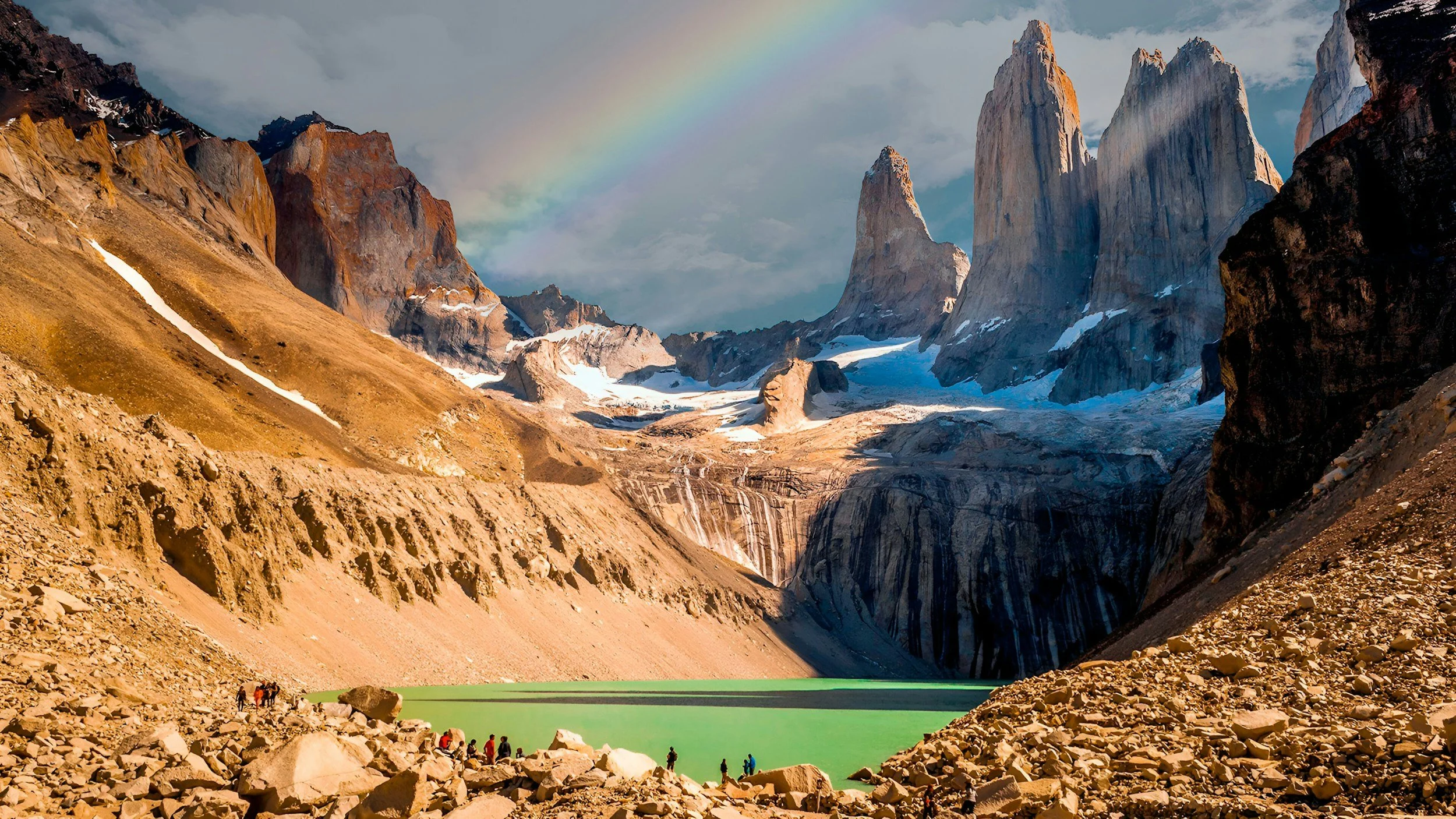 Mountain landscape with rocky terrain, a green lake, and towering snow-capped peaks under a cloudy sky with a rainbow.