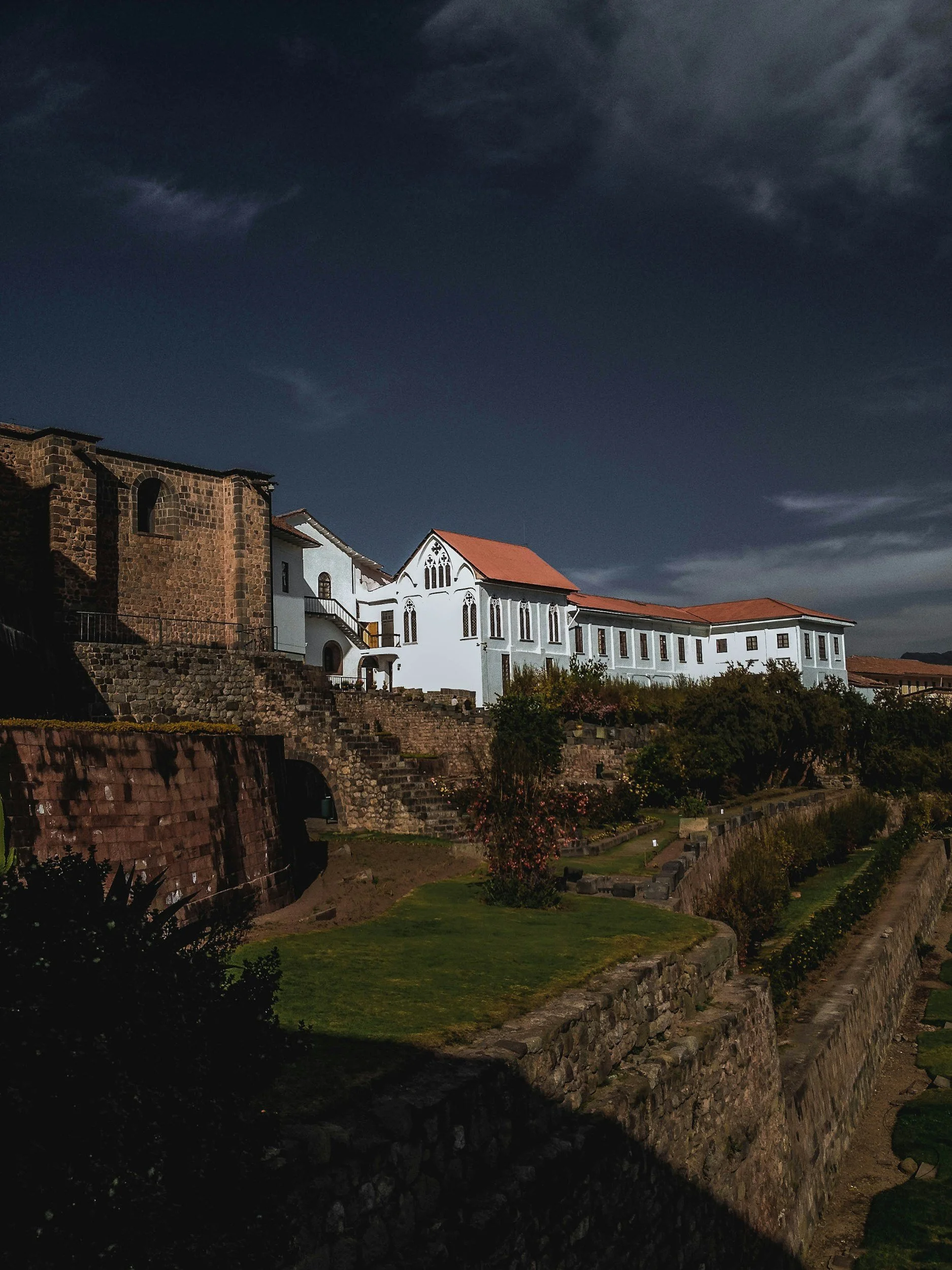A nighttime view of a historic building with white walls and a red-tiled roof, set on a terraced landscape with green gardens, stone walls, and stairs under a dark cloudy sky.