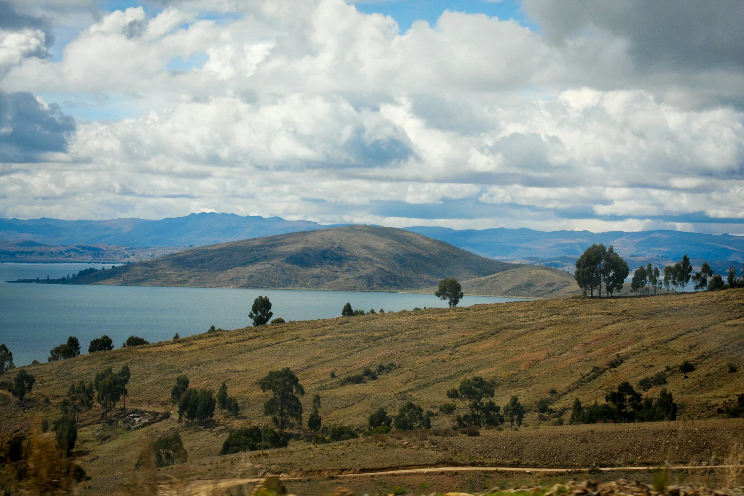 Scenic view of hilly landscape with sparse trees, a lake, and distant mountains under a cloudy sky.