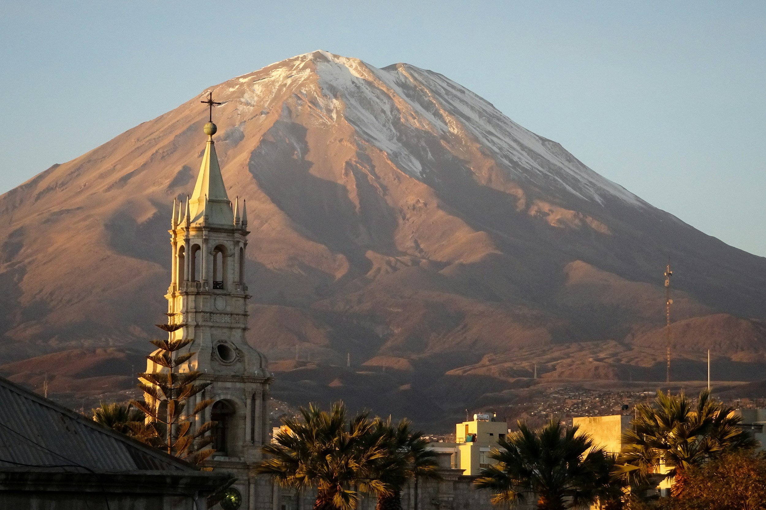 A mountain with snow on its peak in the background, and a church steeple with a cross on top in the foreground, with some palm trees and buildings at the base.