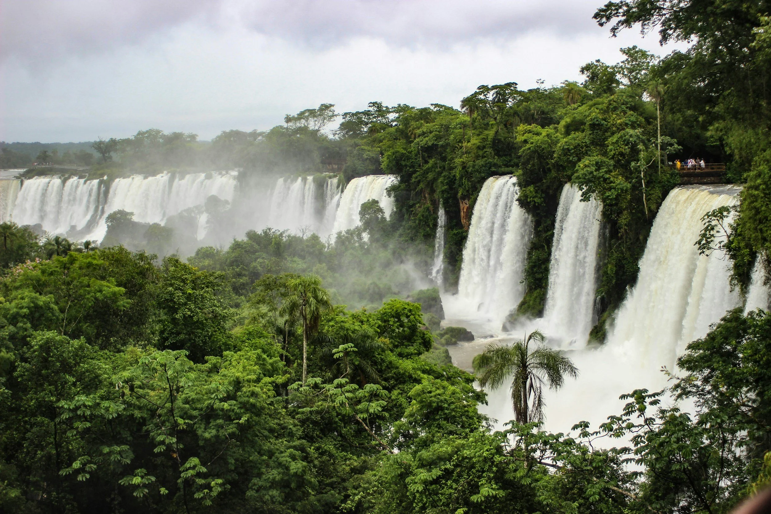 Waterfalls flowing over a lush green rainforest in a misty, cloudy atmosphere, with viewing platforms on the right side and dense trees in the foreground.