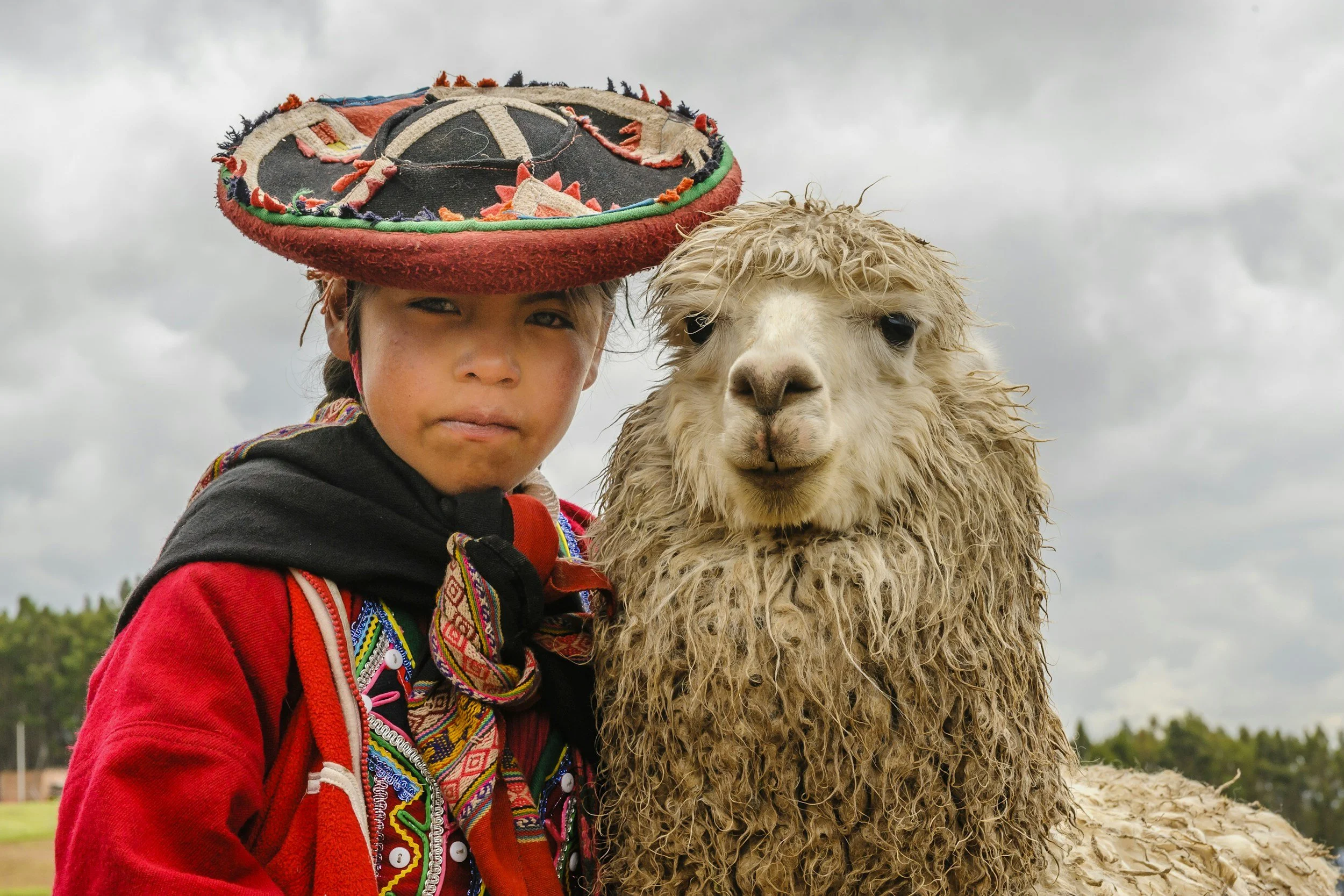 A young person in traditional colorful clothing and a hat with embroidery, standing next to a beige alpaca outdoors on a cloudy day.