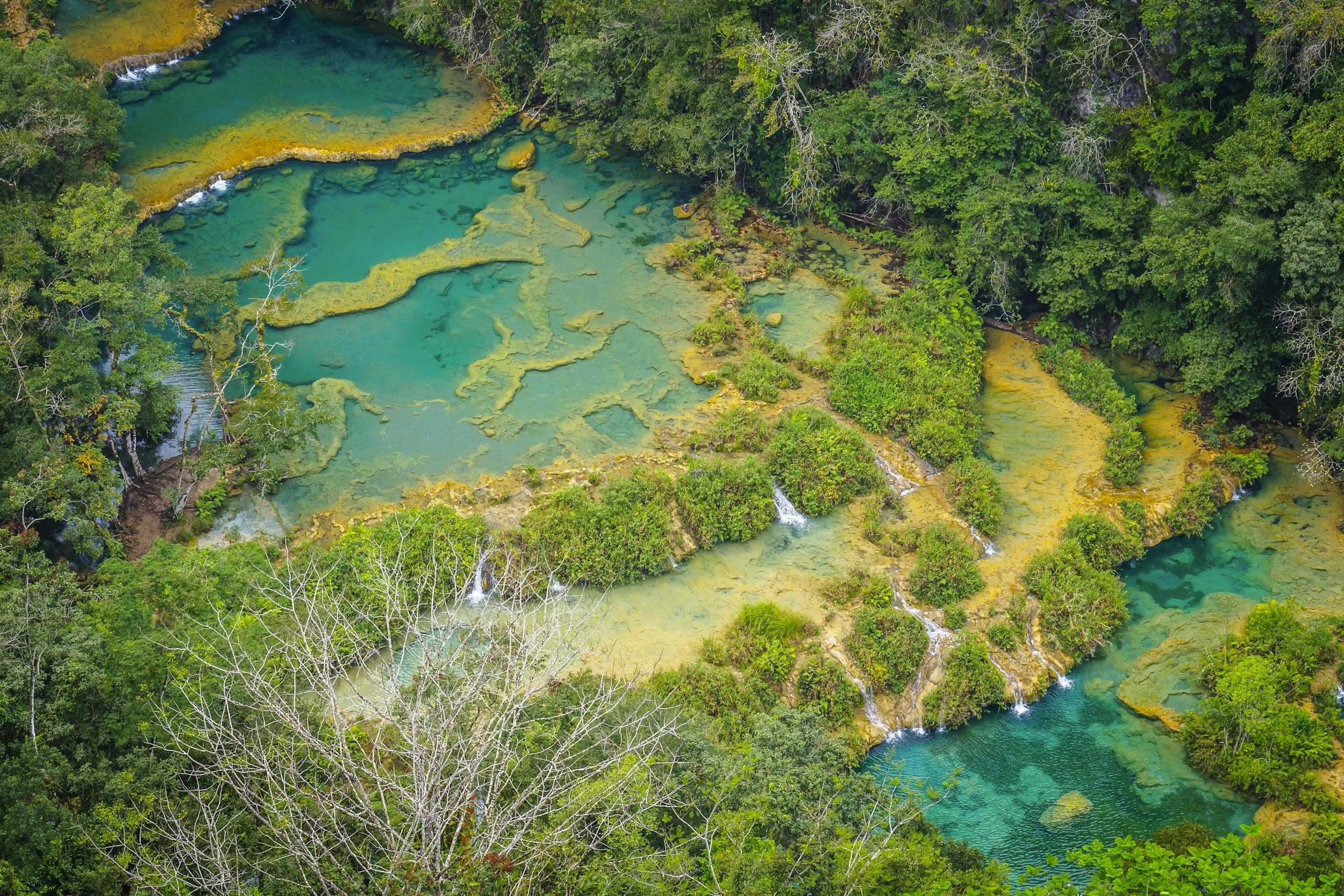 Aerial view of turquoise hot springs with small waterfalls in lush green forest.
