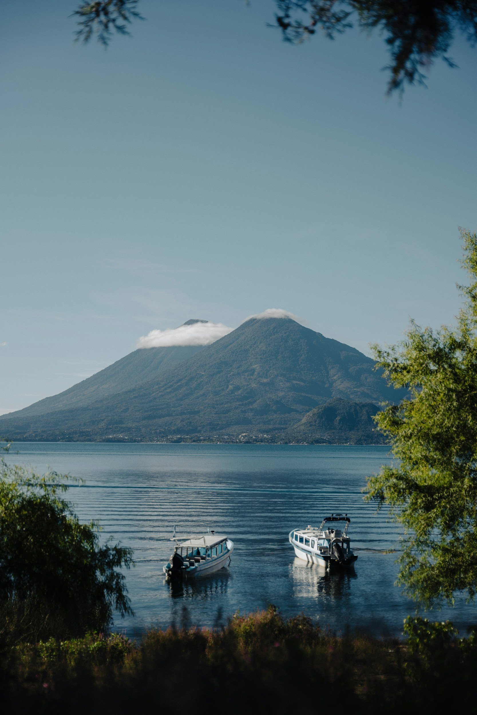 A peaceful lake scene with two boats floating on calm water, a mountain in the background, and trees framing the image.