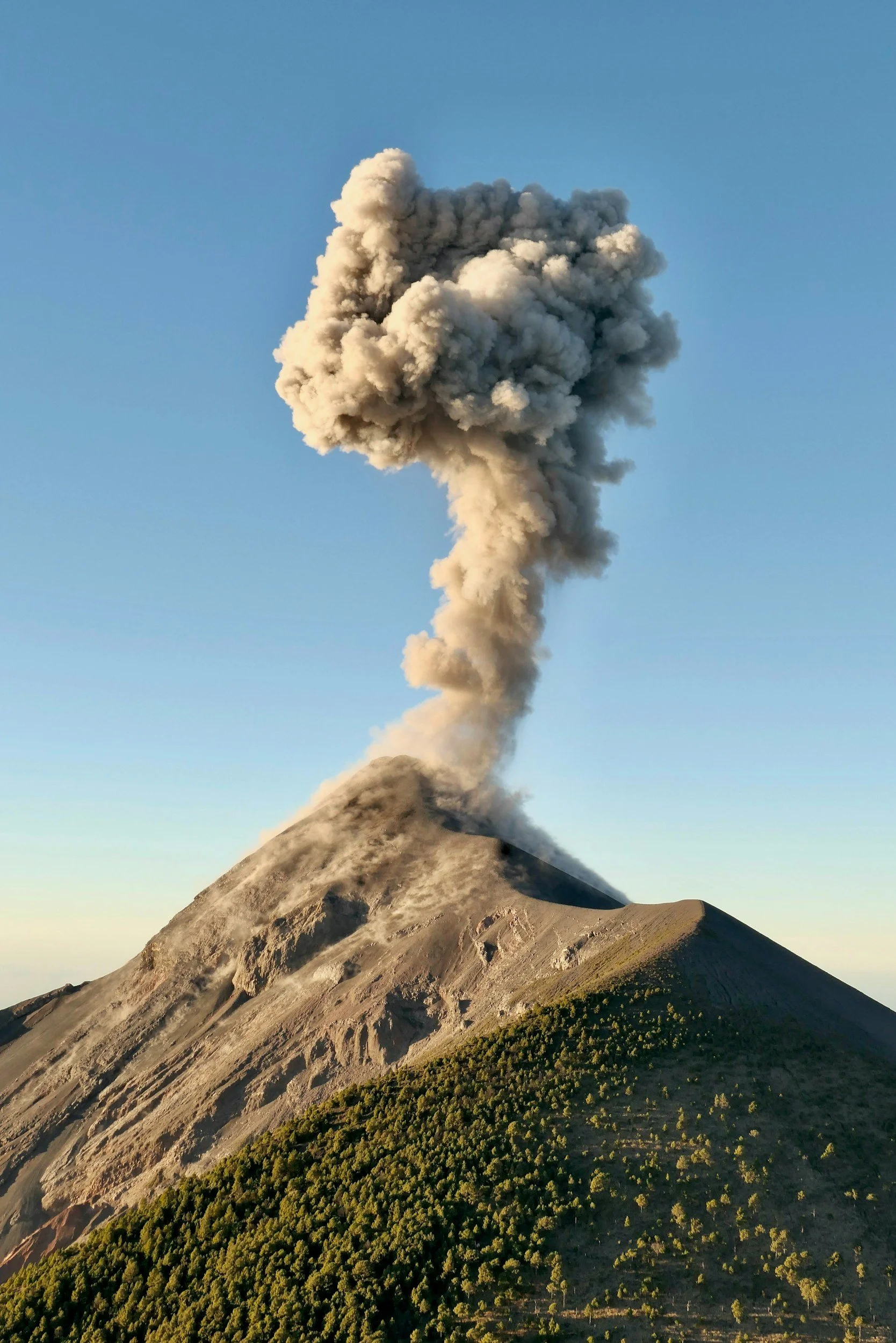 An erupting volcano releasing a large plume of ash and smoke into the sky, with green vegetation on the lower slopes.