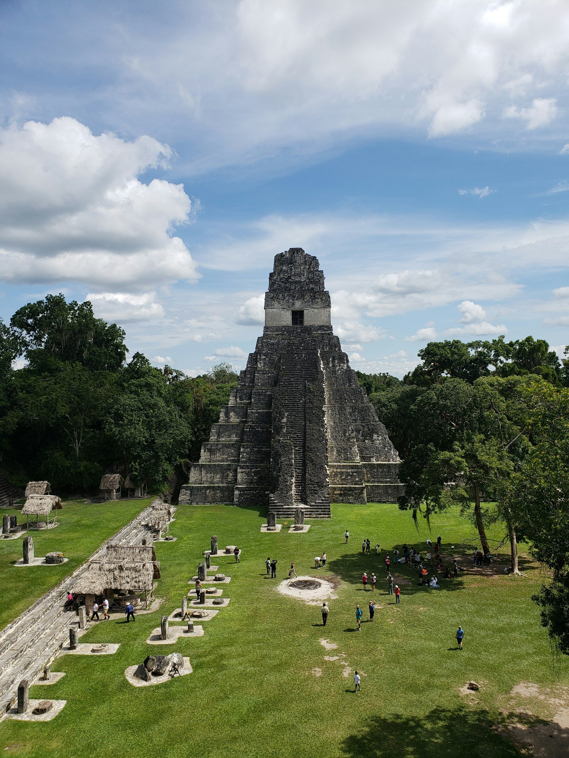 Ancient Mayan pyramid with a stepped structure, surrounded by lush green trees, with tourists exploring the site under a partly cloudy sky.