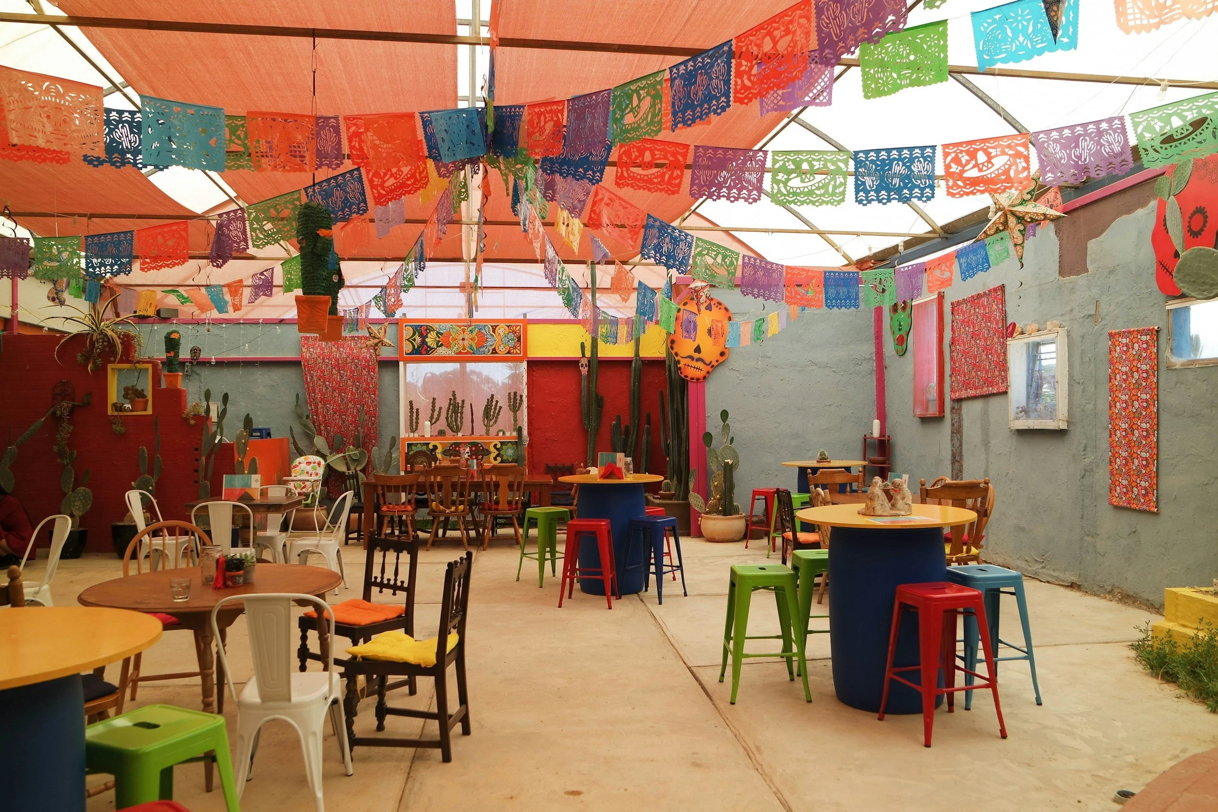 Colorful outdoor patio decorated for a celebration with papel picado banners hanging from the ceiling, cactus plants along the walls, and various tables and chairs in bright colors.