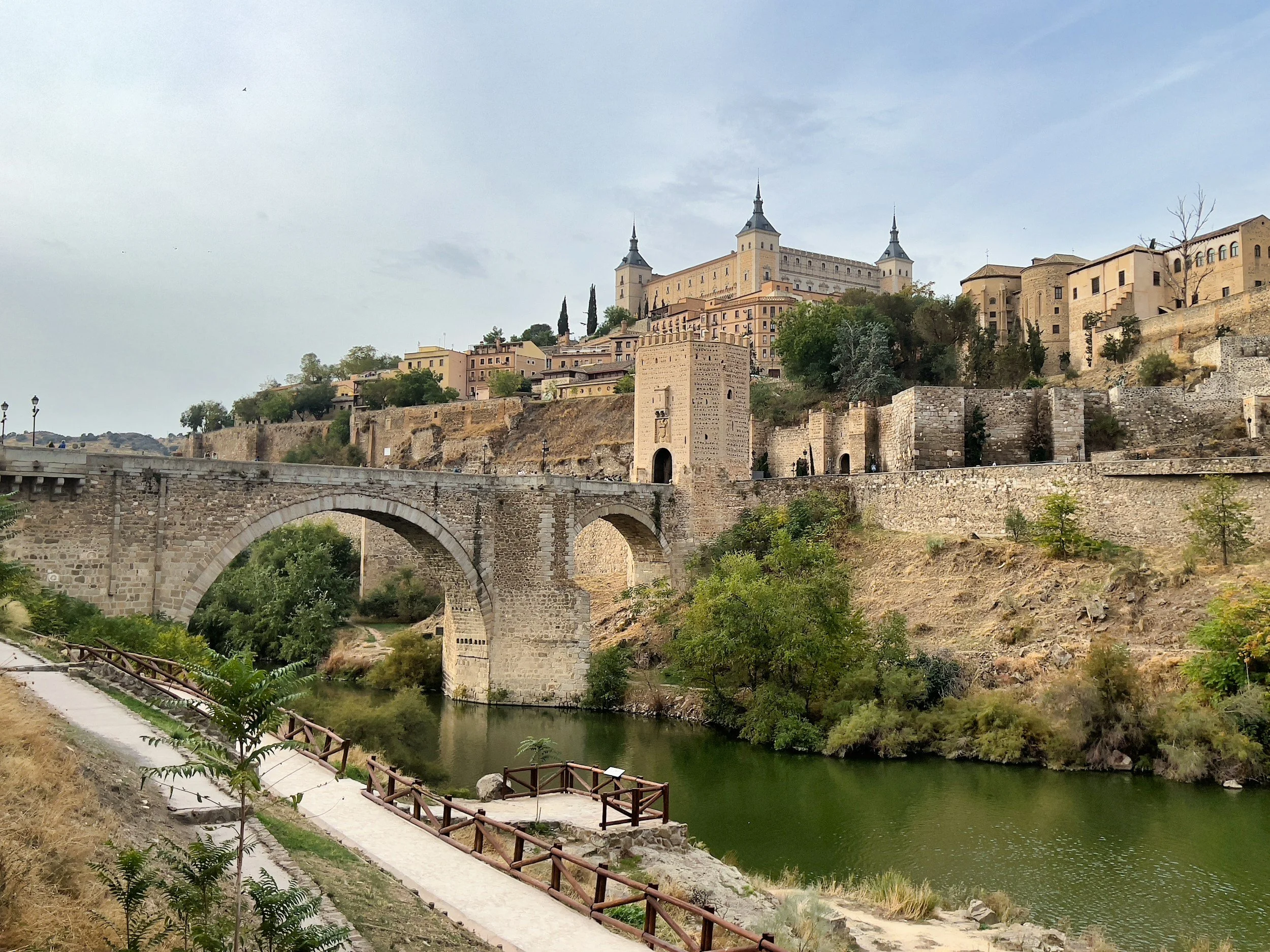 Vista de un castillo en la colina, con una estructura de puente antiguo sobre un río, con árboles y un camino peatonal en primer plano.