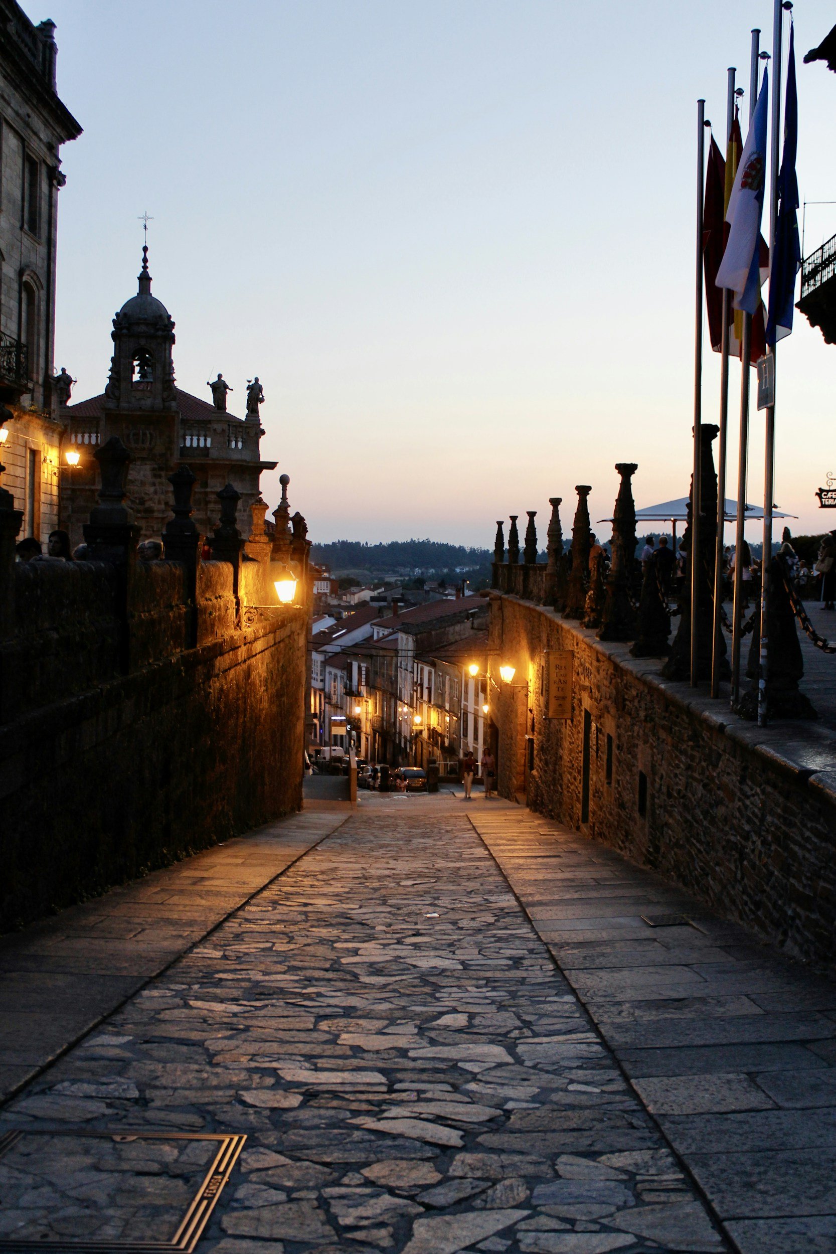 Calles empedradas de un pueblo durante el atardecer, con casas antiguas, farolas encendidas y banderas ondeando.