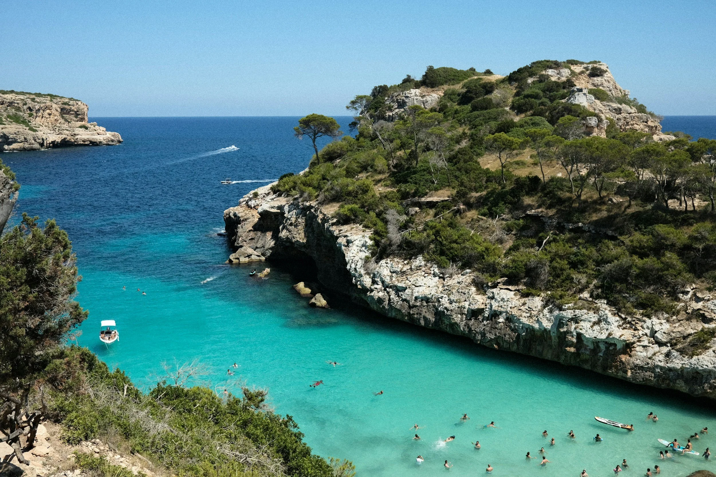 Paseo por una cala con agua cristalina y rodeada de rocas y vegetación, con personas nadando y un bote en el agua.