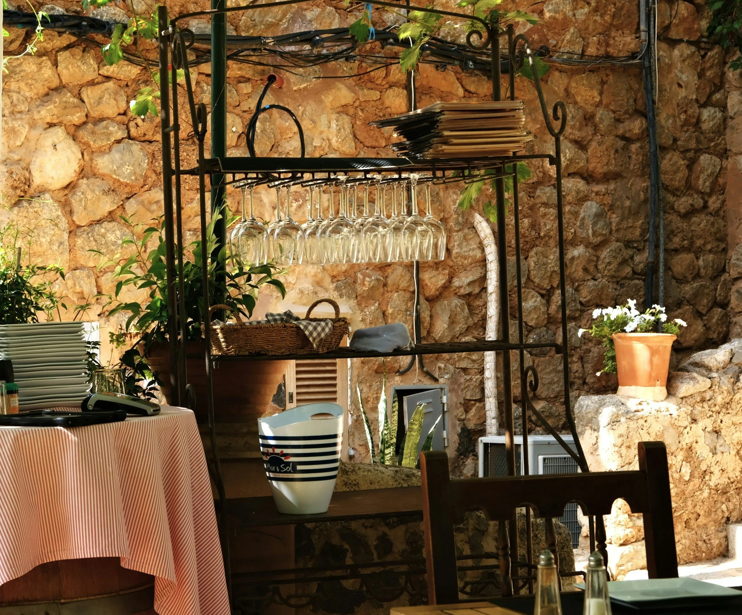 Una estantería de hierro negro con copas de vino colgadas, platos apilados, y algunos papeles, en un patio con muro de piedra y plantas.