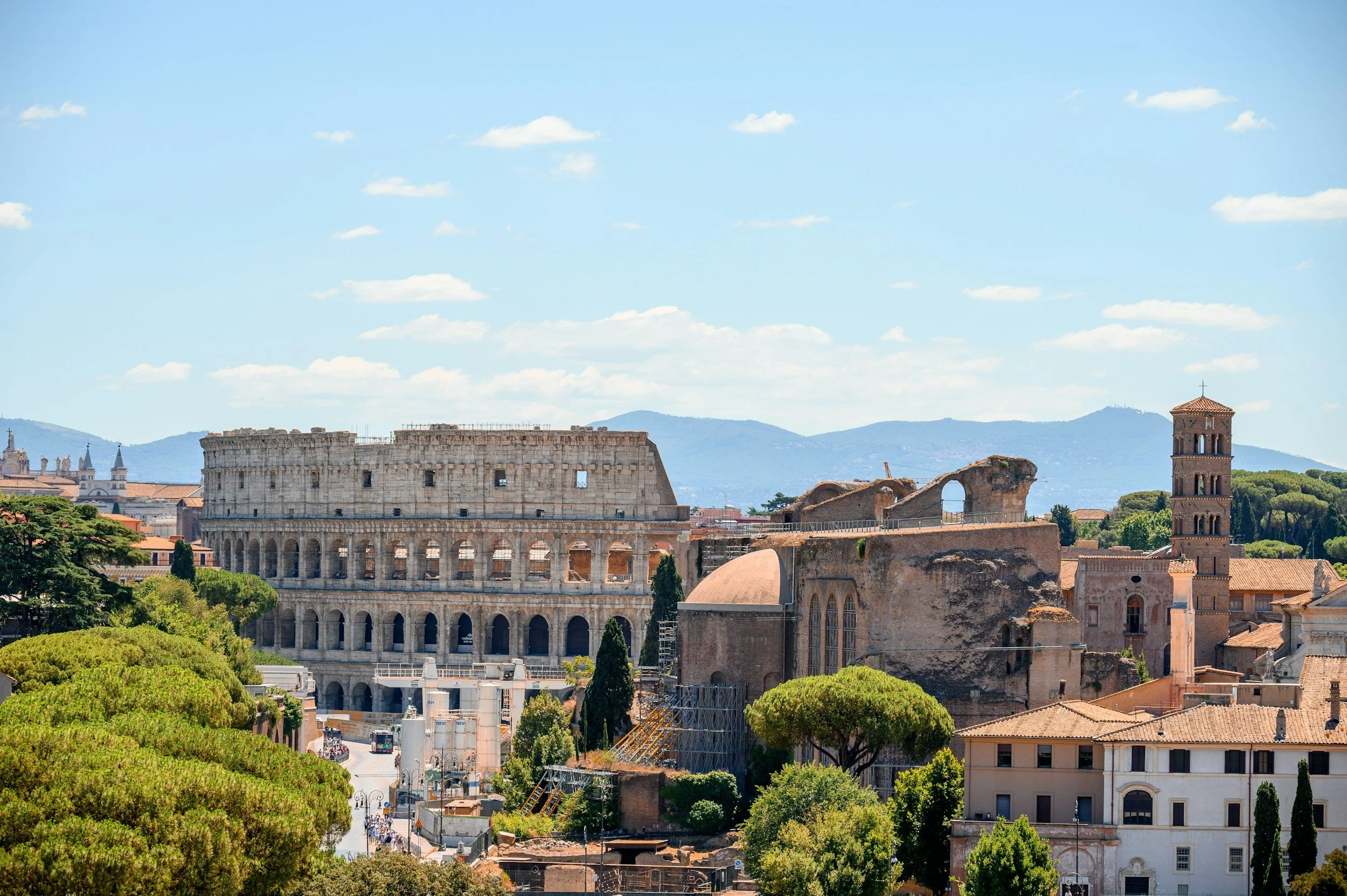 Vista del Coliseo Romano y otras estructuras históricas en Roma, Italia, con árboles verdes y montañas en el fondo bajo un cielo azul con nubes blancas.