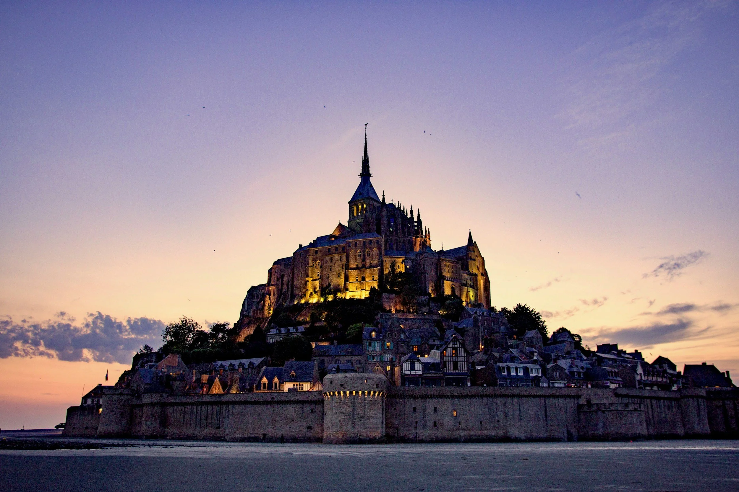 Mont Saint-Michel iluminado durante el atardecer, visto desde la playa con el cielo en colores suaves.