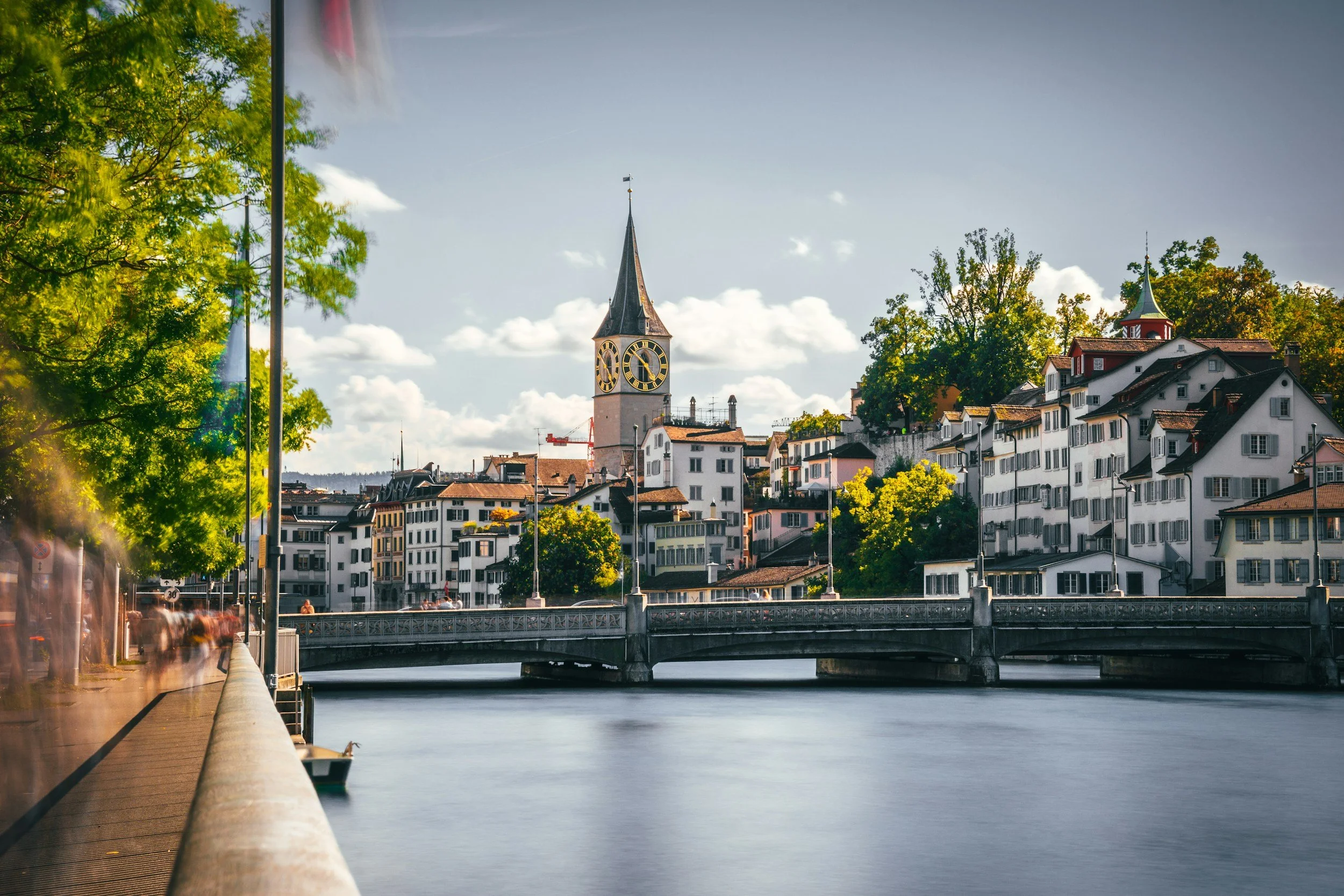 Paisaje de un río con un puente, edificios blancos y una iglesia con reloj en una ciudad rodeada de árboles y montañas al fondo en un día soleado.