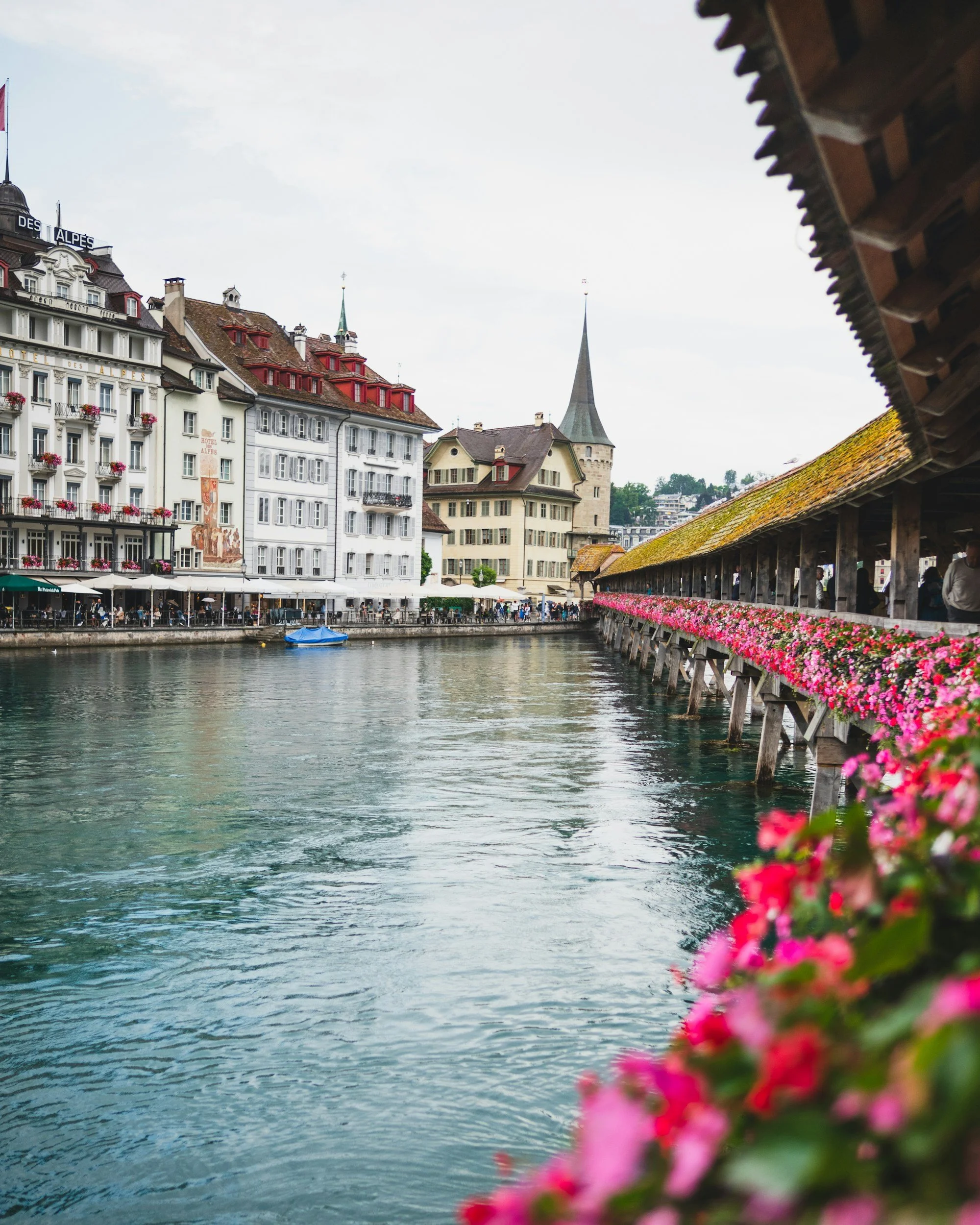 Vista de un río en una ciudad europea con edificios históricos y un muelle adornado con flores. Se puede ver una iglesia con una torre alta en el fondo y un pequeño barco azul en el río.
