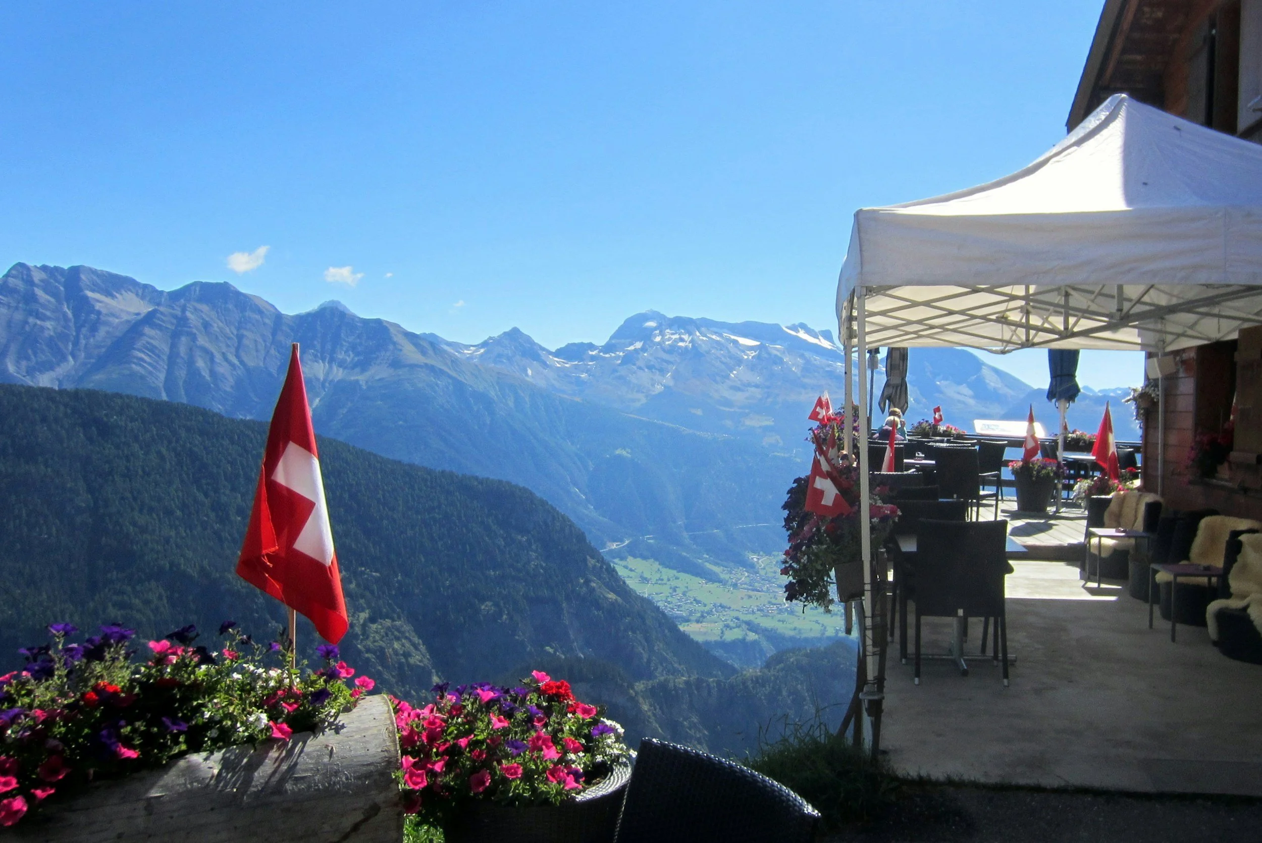 Terraza de un restaurante con vista a las montañas nevadas en Suiza, decorada con banderas suizas y flores en macetas, en un día soleado.