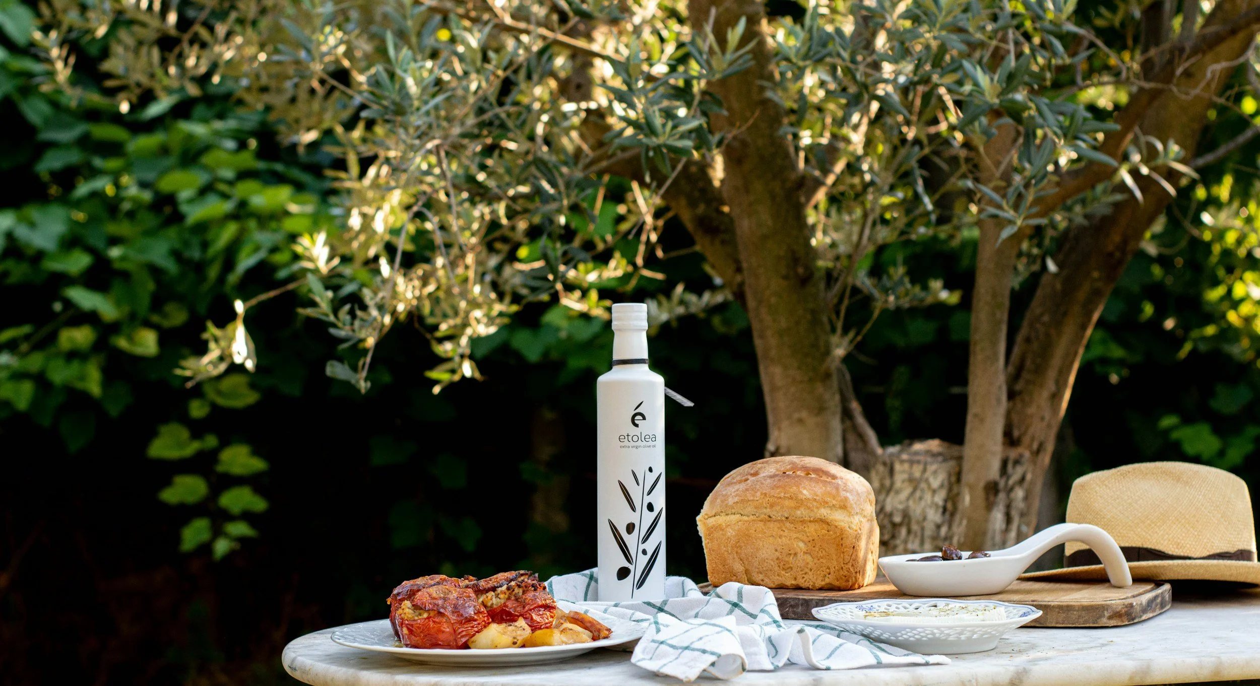 Mesa con pan, comida, botella de aceite de oliva y sombrero en un entorno al aire libre con árboles y vegetación.