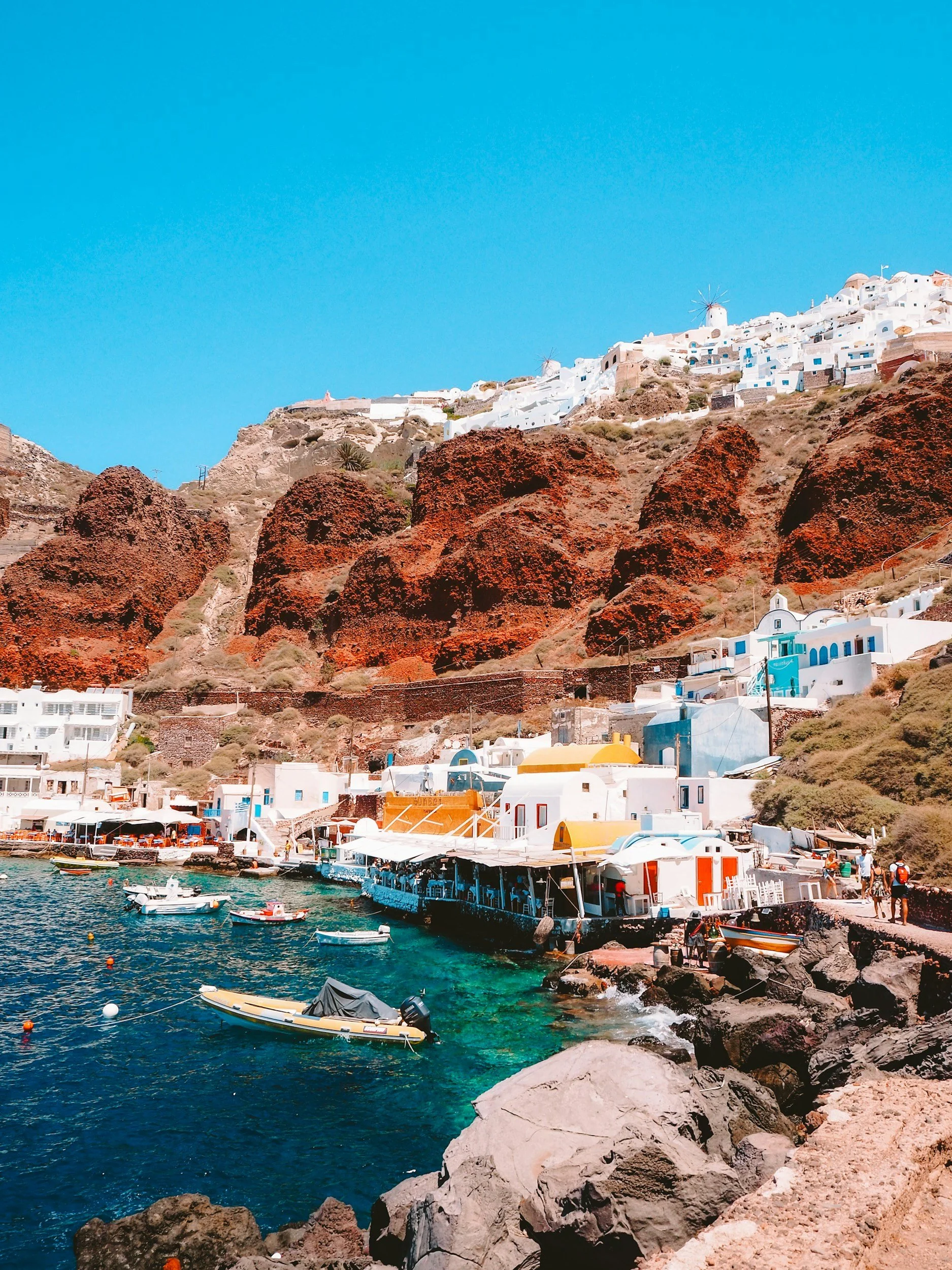 Paisaje marítimo en un pueblo costero con casas blancas y molinos en una colina, cerca del mar con barcos en el agua y un cielo despejado.