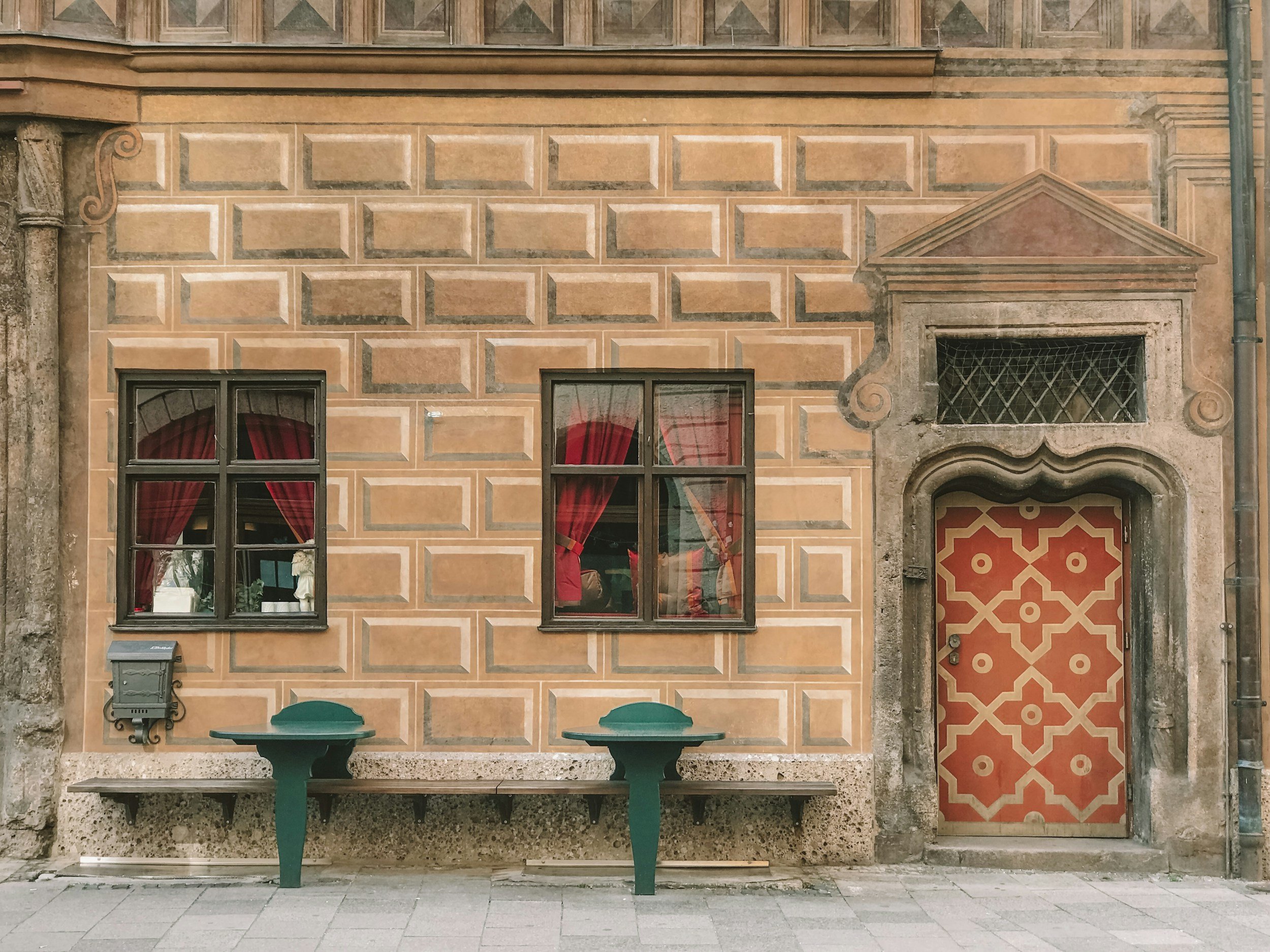 fachada de un edificio con paredes de ladrillos en tonos tierra, ventanas con cortinas rojas, un buzón metálico y un banco. Puerta con diseño geométrico en tonos rojos y puertas de ventanas con rejas de protección.