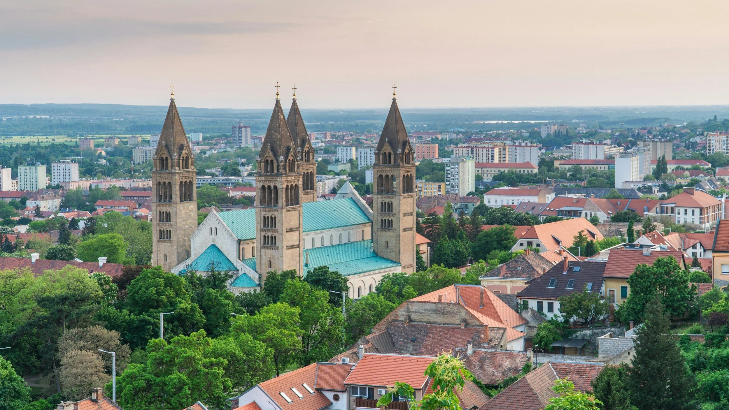 Vista aérea de una ciudad con una iglesia gótica de tres torres en primer plano y edificios residenciales y comerciales en el fondo, rodeados de árboles verdes.