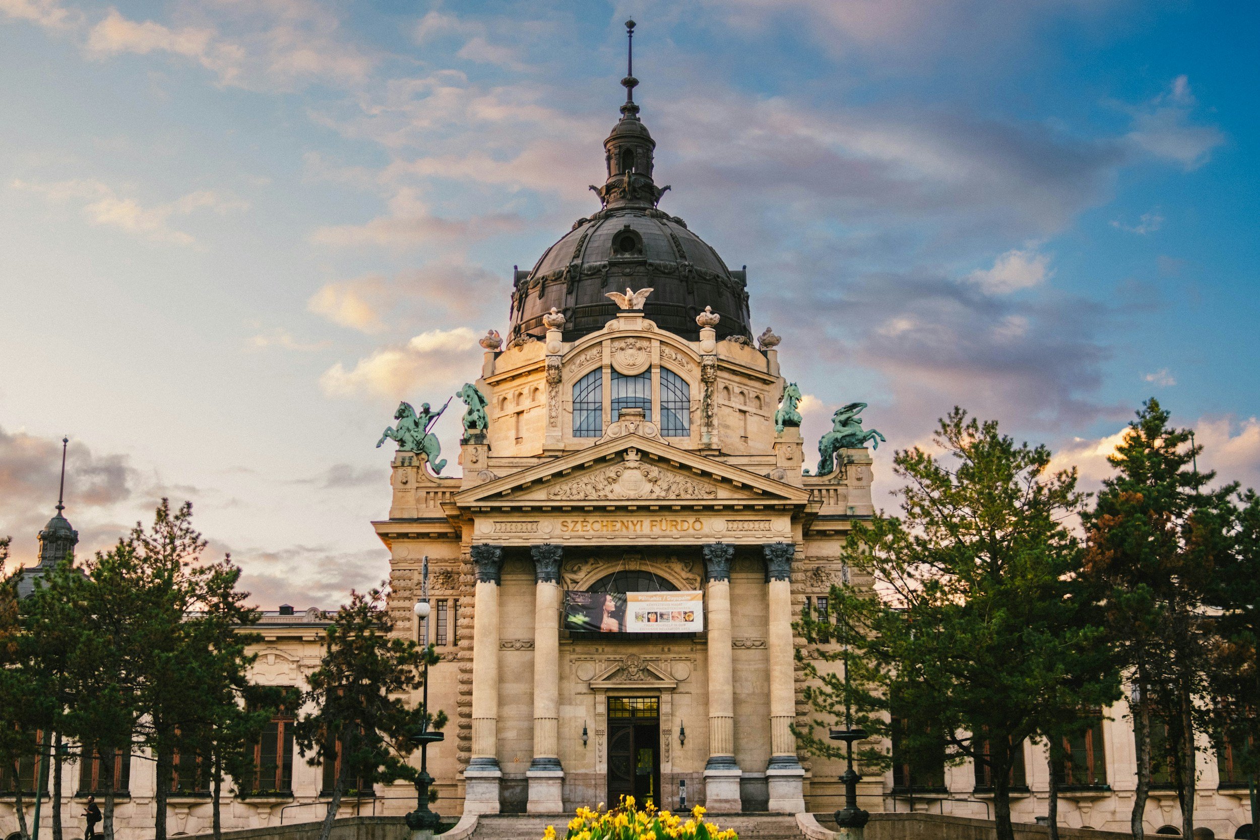 Edificio histórico con cúpula y estatuas en la azotea, rodeado de árboles y cielo al atardecer.