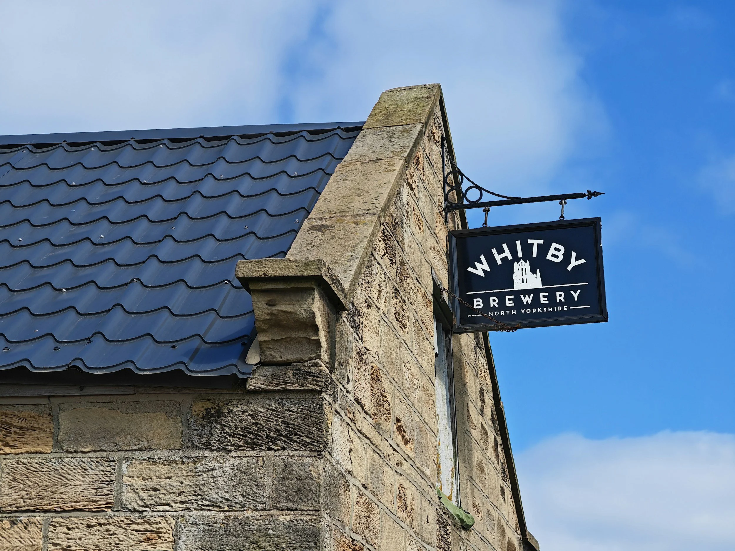 Letrero de la cervecería Whitby en un edificio de piedra con tejado de tejas azules, bajo un cielo nublado y azul