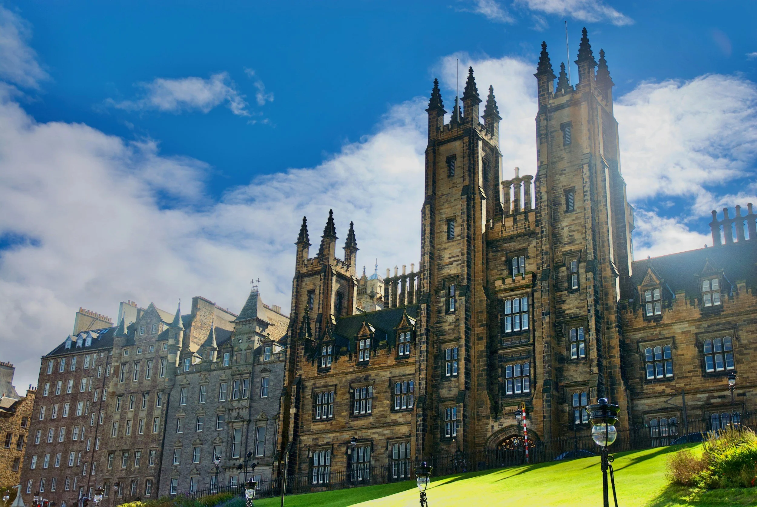 Castillo antiguo de piedra con torres altas y ventanas grandes, bajo un cielo azul con nubes, rodeado de césped y farolas en primer plano.