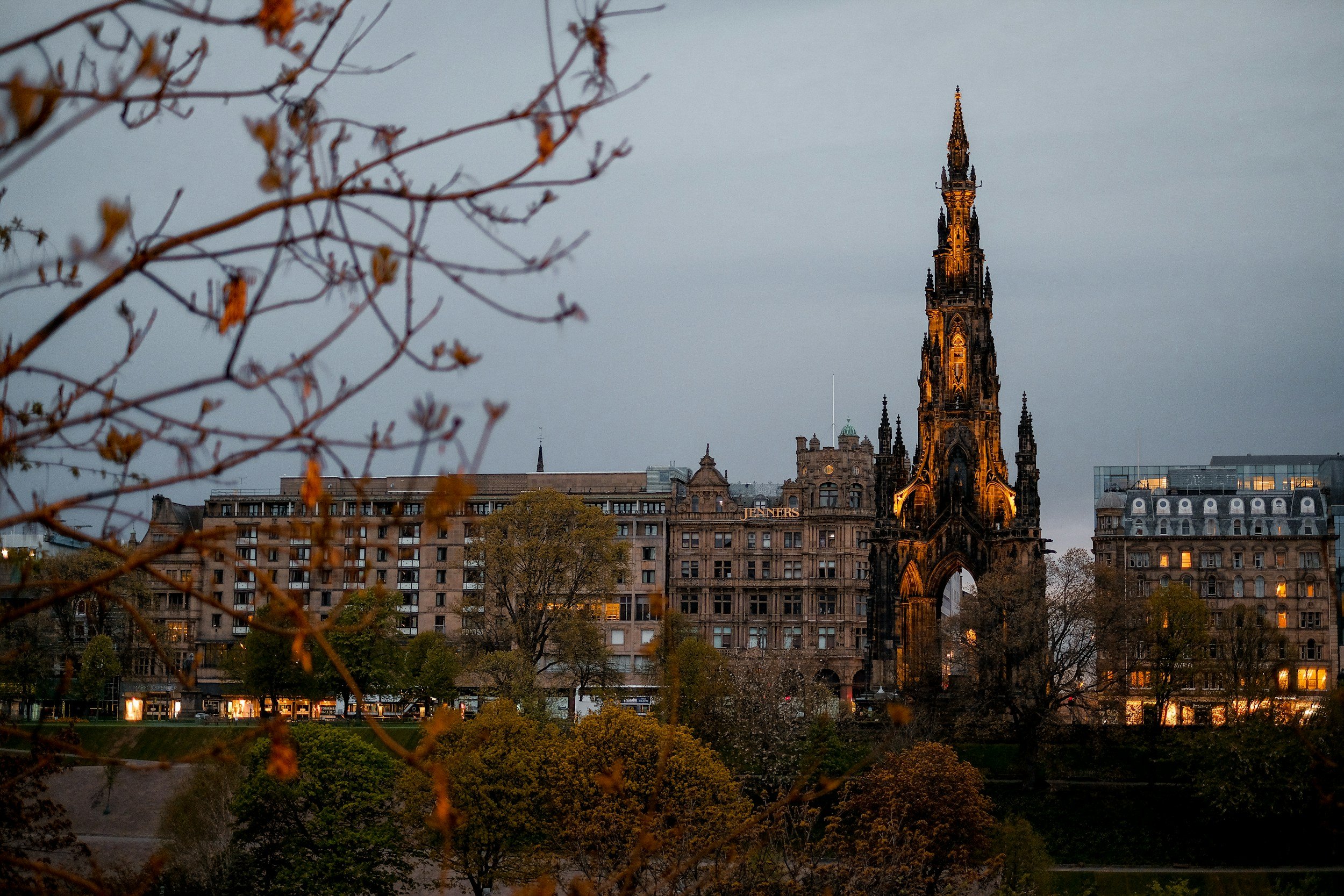 Vista del puente de Martín Lutero en Edimburgo, con un monumento histórico iluminado y árboles en primer plano en un atardecer nublado.