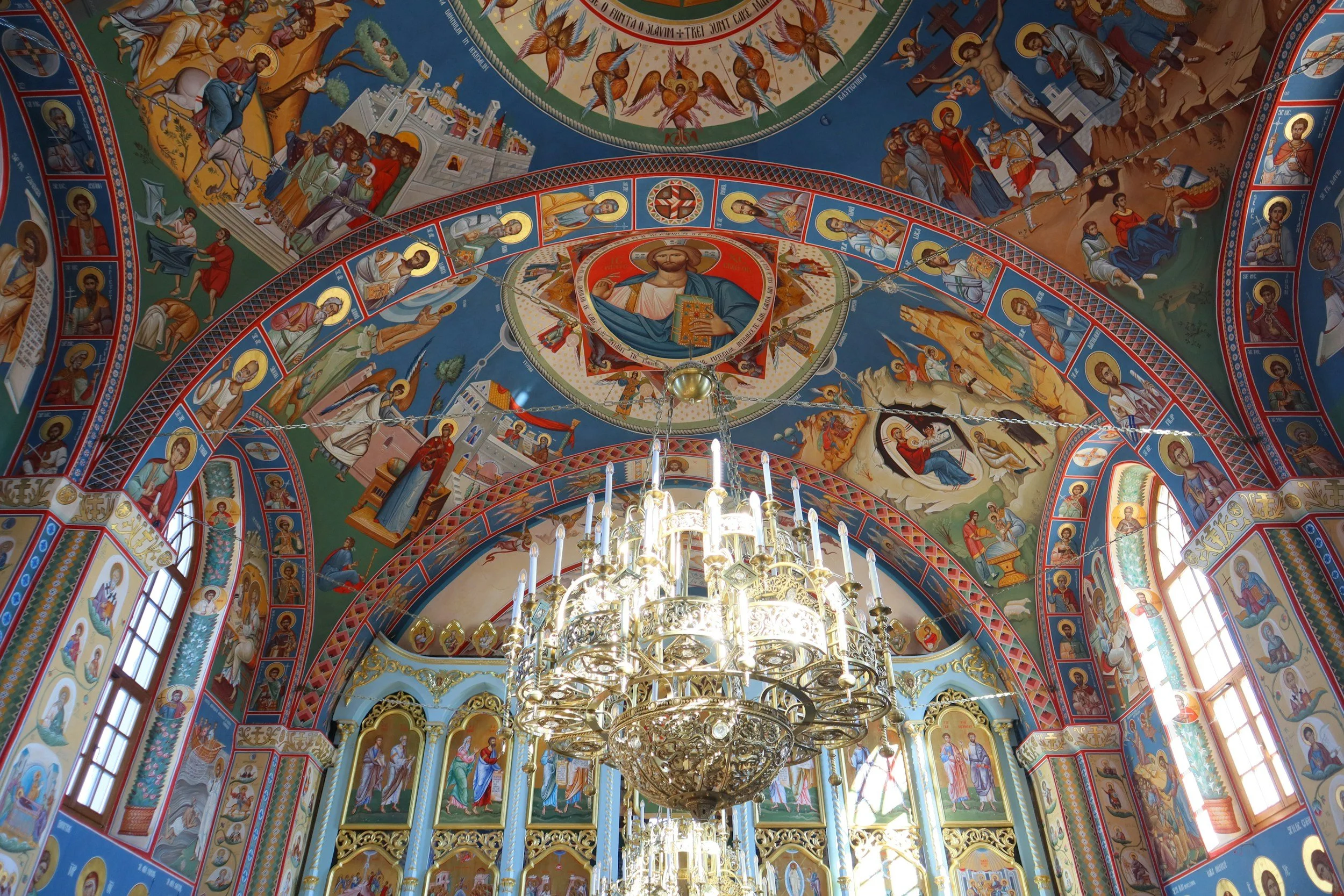 Interior de una iglesia ortodoxa con frescos religiosos en las paredes y el techo, y un candelabro grande de oro colgado en el centro.