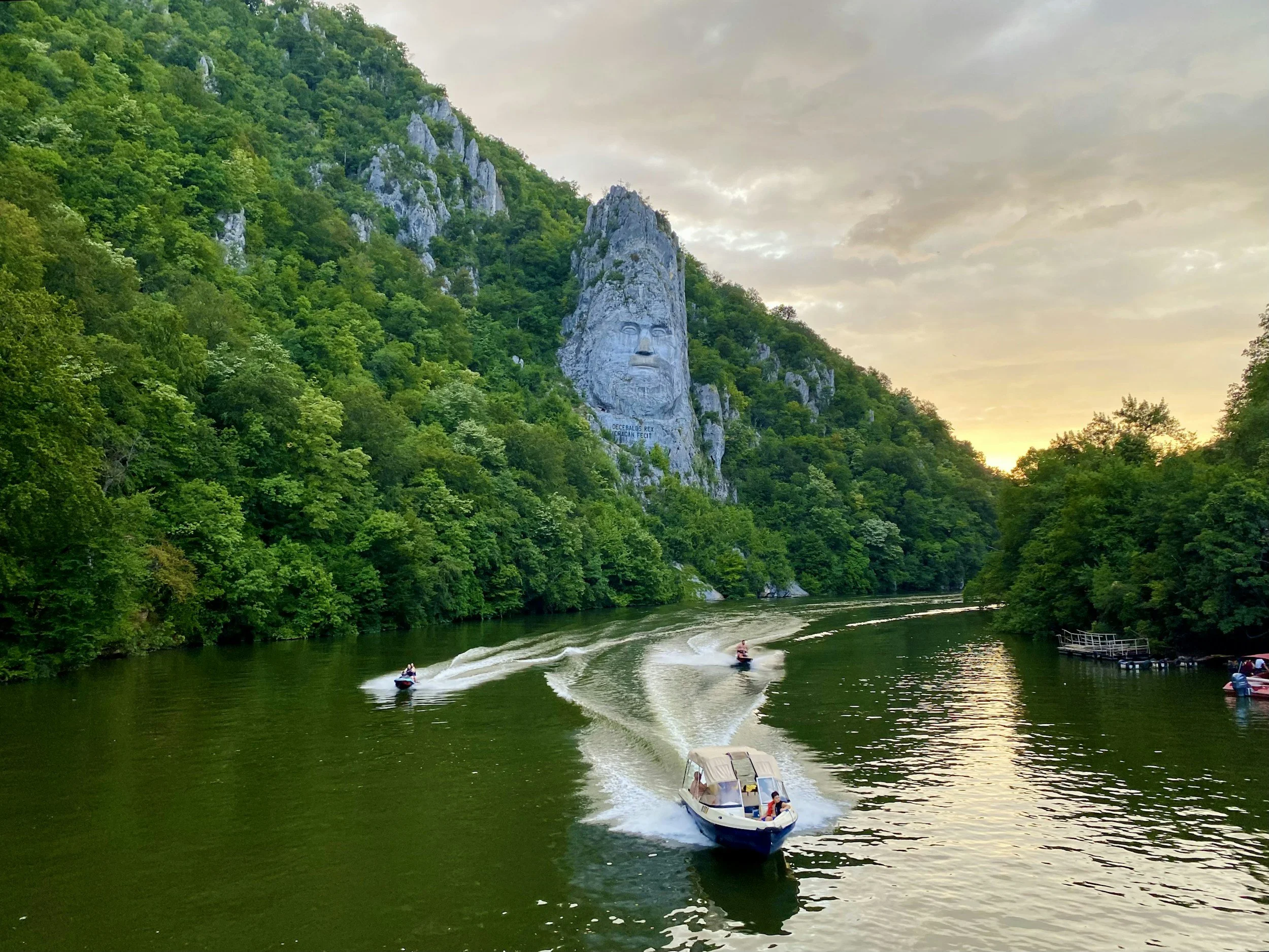 Vista del río con varias personas en lanchas y jet skis, rodeado de jungla y montañas, con una roca gigante con un rostro tallado.