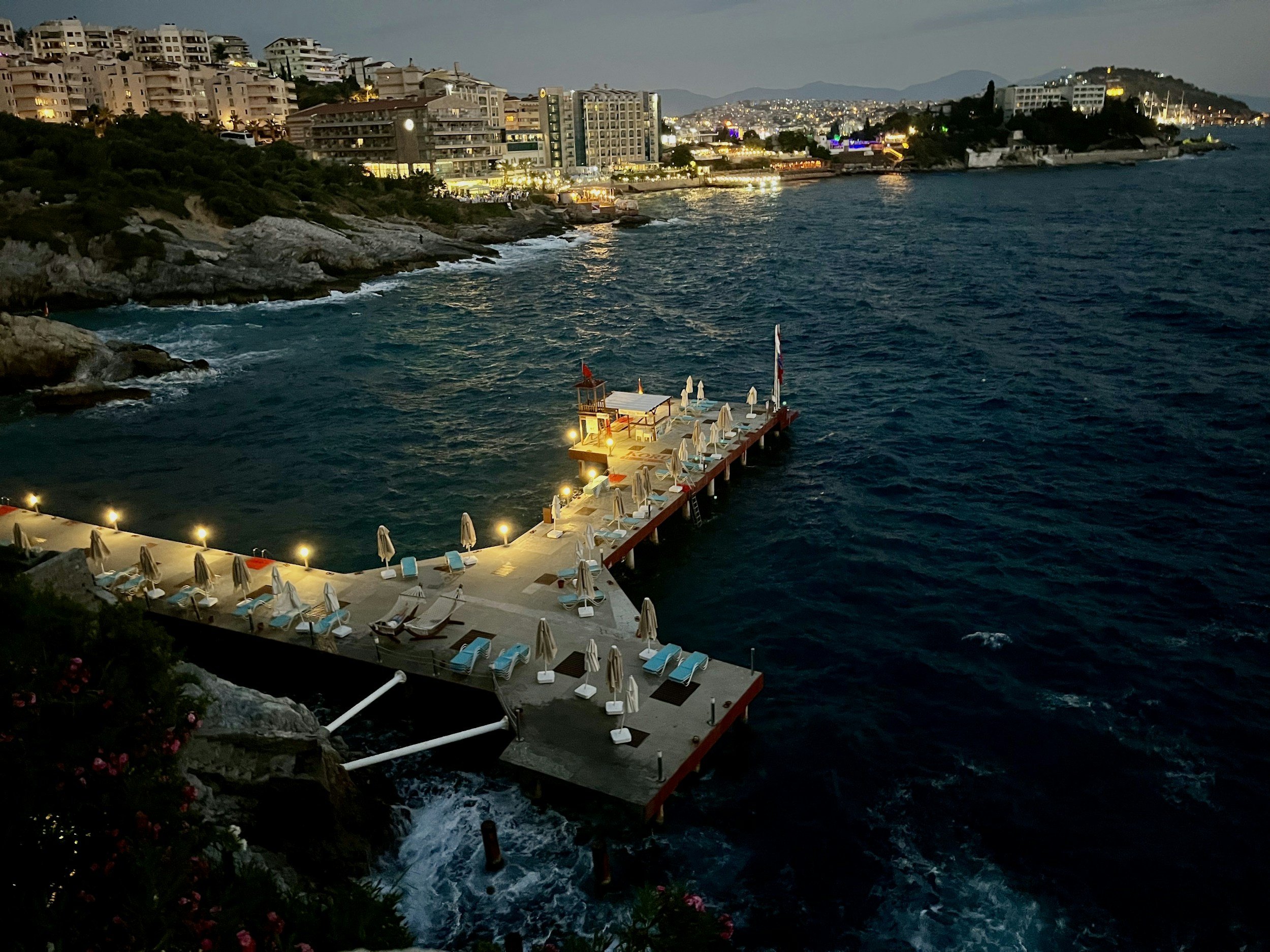 Puerta de muelle iluminada con sillas y sombrillas, vista del mar en la noche y ciudad con edificios iluminados al fondo.