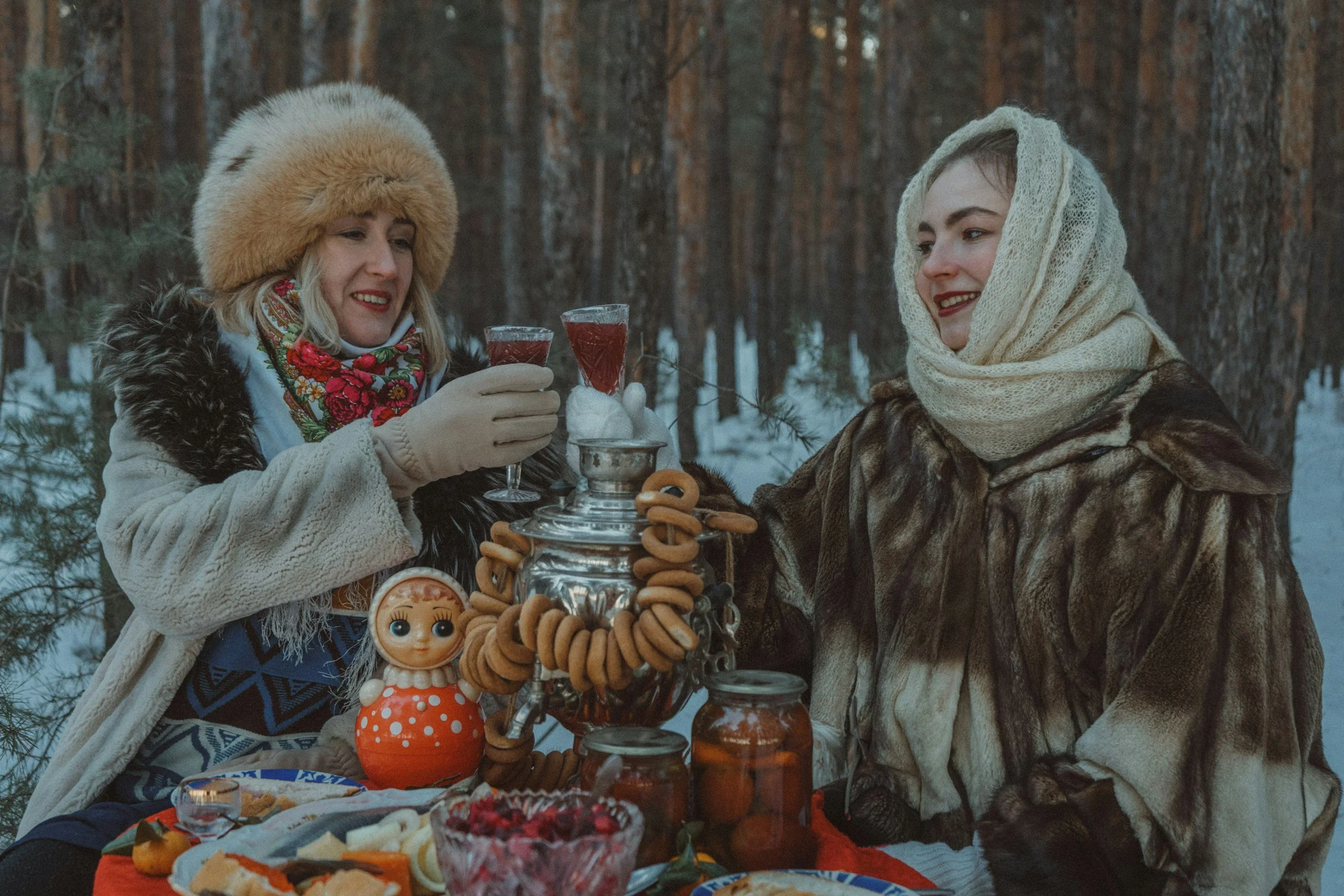 Dos mujeres en un bosque nevado brindando con bebidas calientes, vestidas con ropa de abrigo tradicional rusa, con comida y decoraciones típicas sobre una mesa.